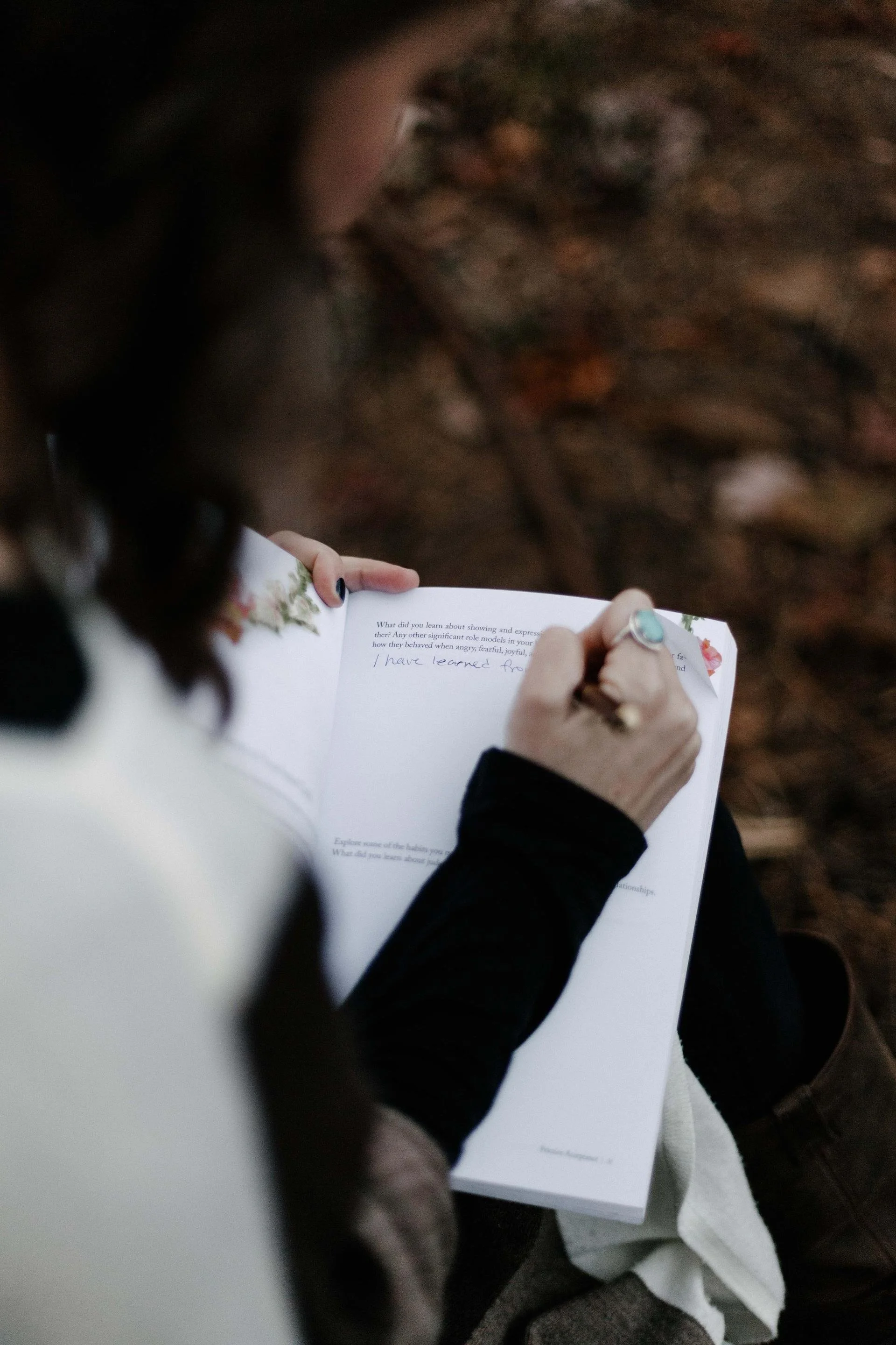 Person sitting outdoors, writing in a notebook with a pen, on a ground covered with leaves and small twigs.