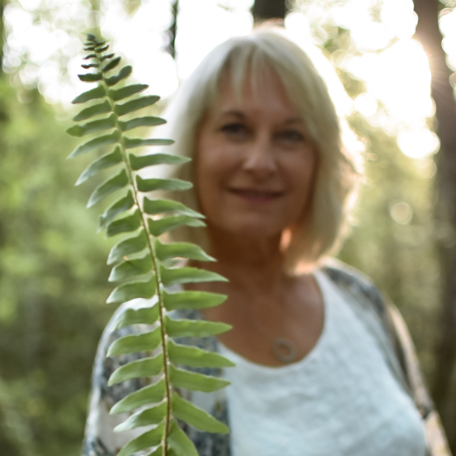 A woman with blonde hair smiling outdoors in a forest, with a fern leaf in the foreground.