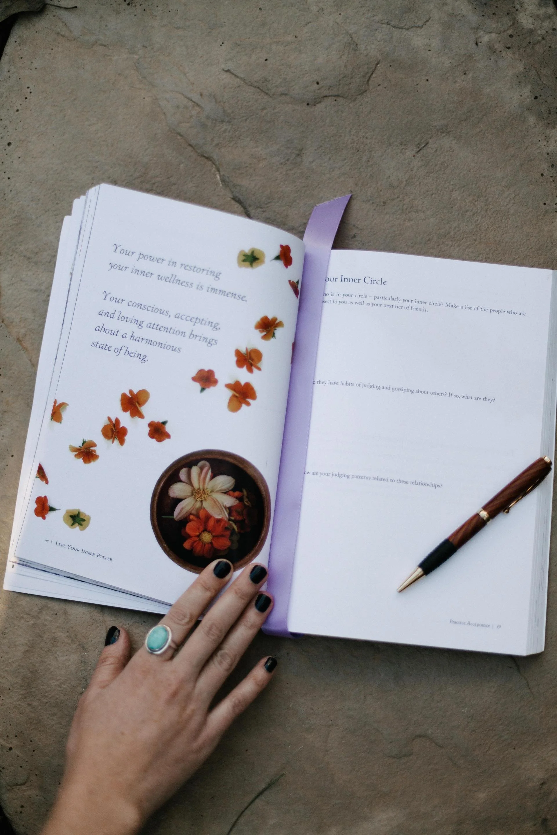 Open book with a purple bookmark on a stone surface, featuring a page with floral illustrations and overlays. A person with dark nail polish and a turquoise ring is holding the book, with a pen resting on the right page.