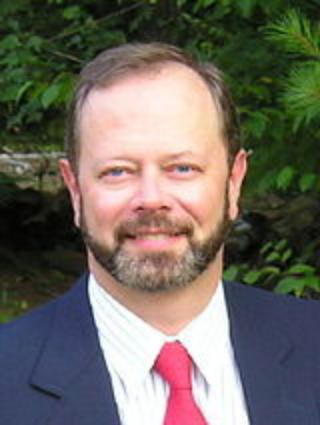 A man with light skin, a beard, and short hair, wearing a navy blazer, white shirt, and red tie, smiling outdoors with green foliage in the background.
