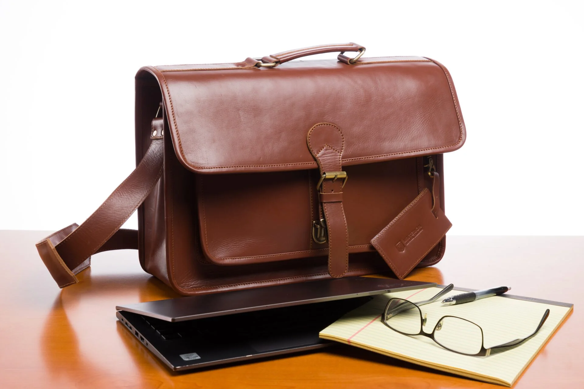 Brown leather briefcase on a wooden desk with a laptop, yellow notepad, pen, and eyeglasses.