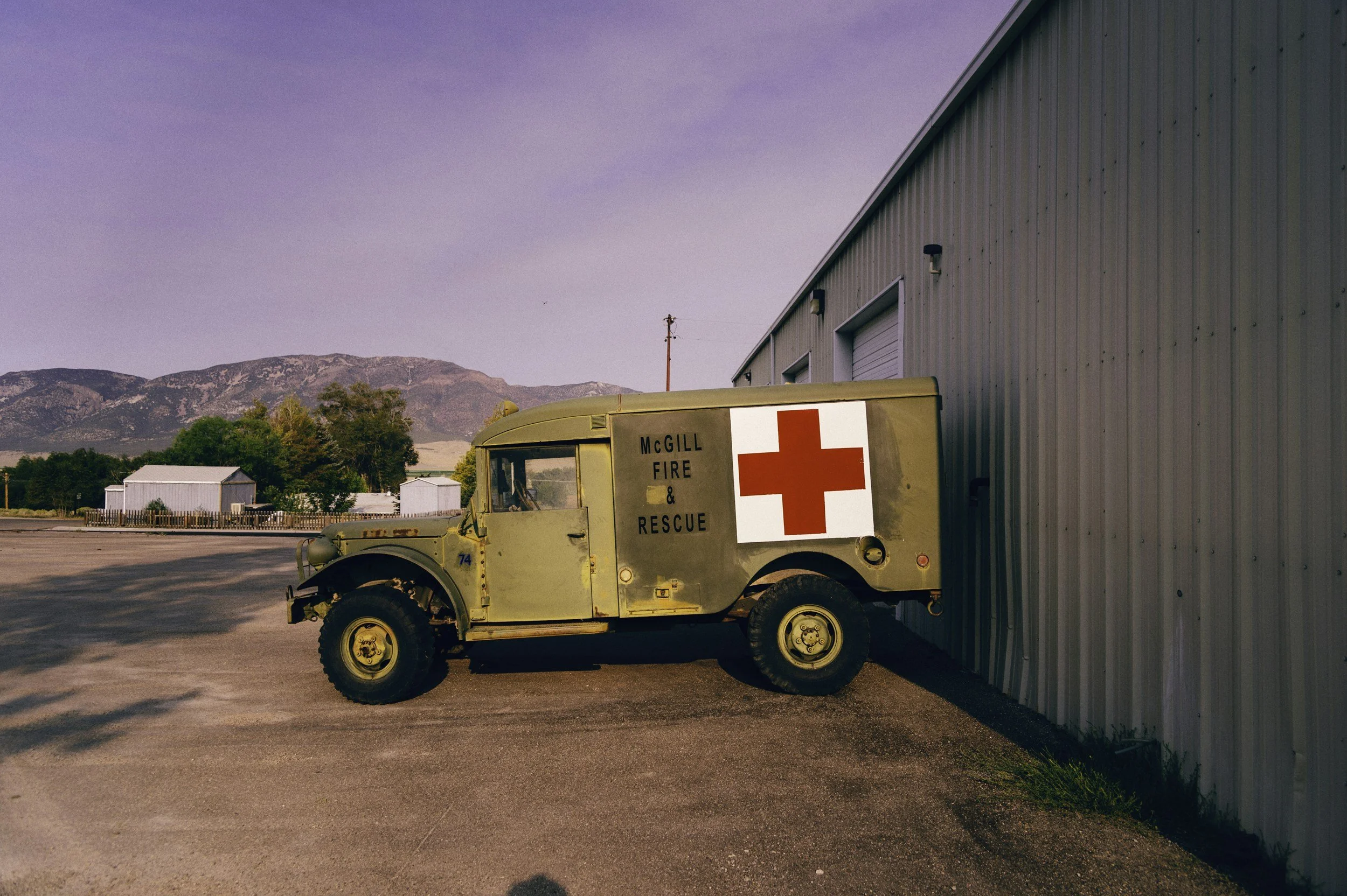 Vintage green fire rescue vehicle with a red cross symbol and 'McGill Fire & Rescue' written on its side, parked beside a large metal building in a rural setting with mountains in the background.