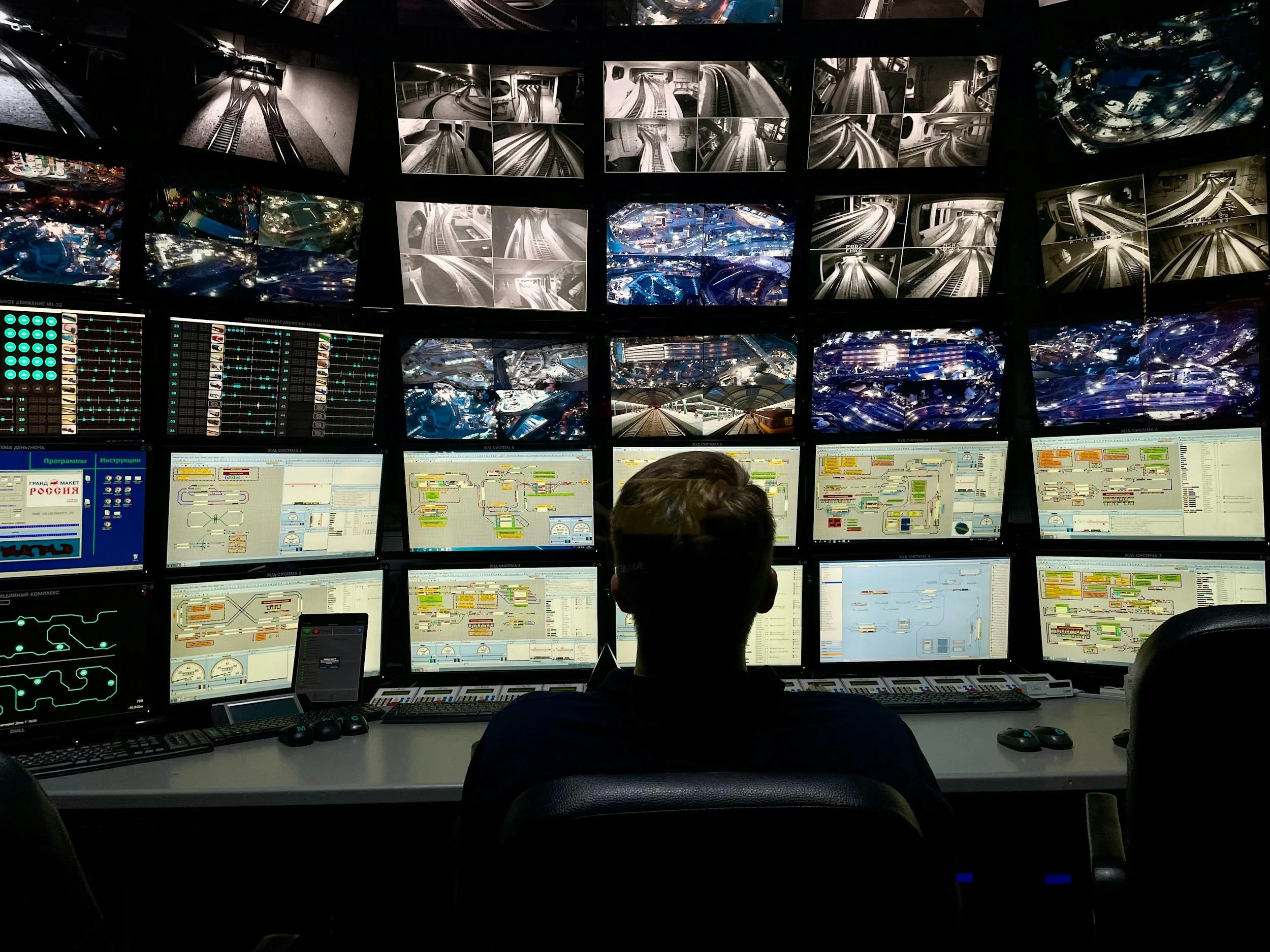 A person sitting in front of multiple monitors displaying train and railway control systems, including surveillance footage of tracks and signals.