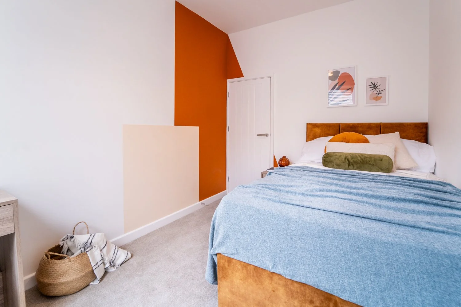 A bedroom with white walls, a wooden bed with a light brown headboard, blue bedspread, and various pillows including an orange one and a green bolster. Two framed art prints hang above the bed. To the left, an orange accent wall and a small nightstand with an orange vase. A closed white door is in the corner, and a woven basket with a striped blanket on the beige carpet near the wall.