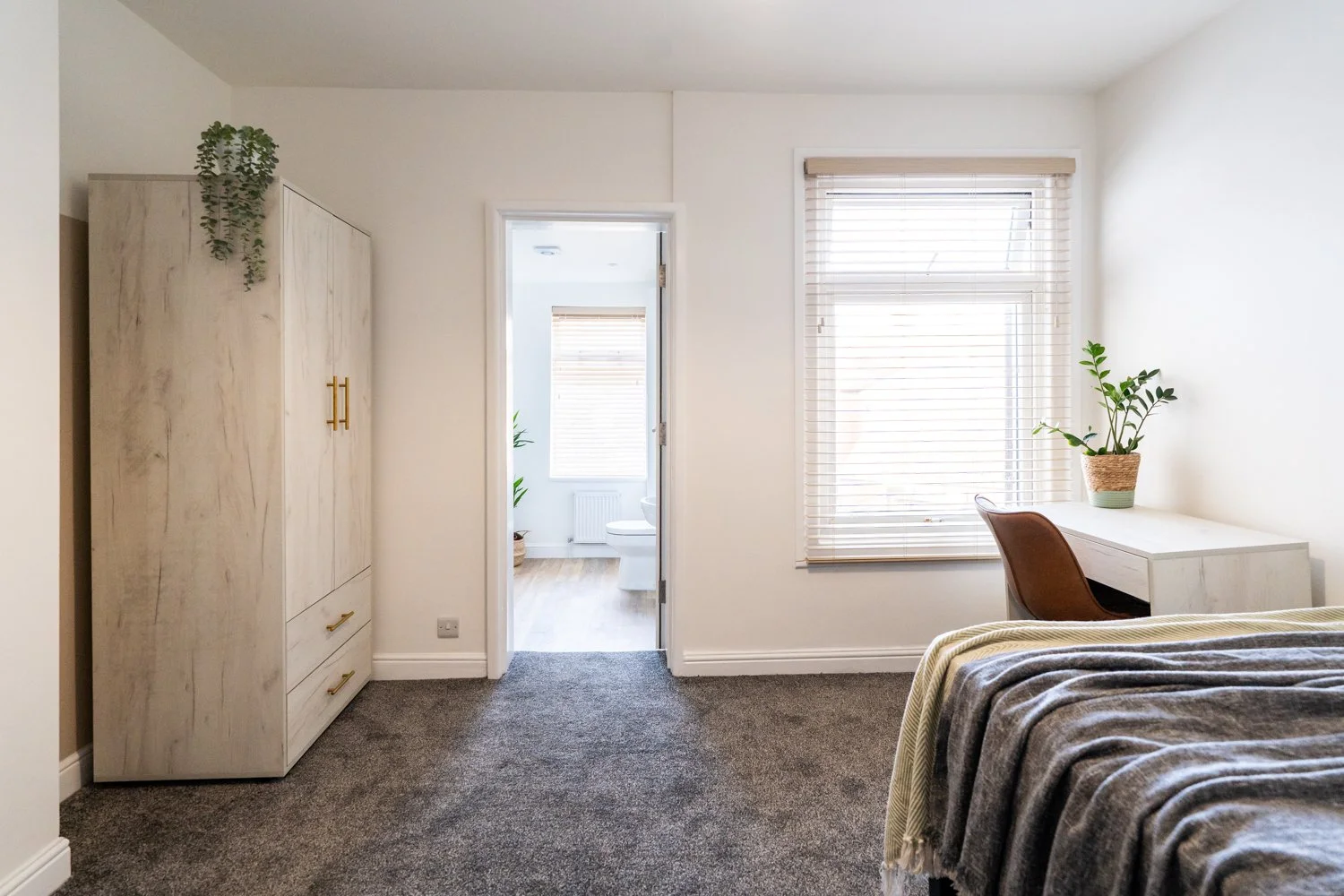 Bright bedroom with a window, wooden desk with potted plant, wardrobe, and bed with gray blanket.
