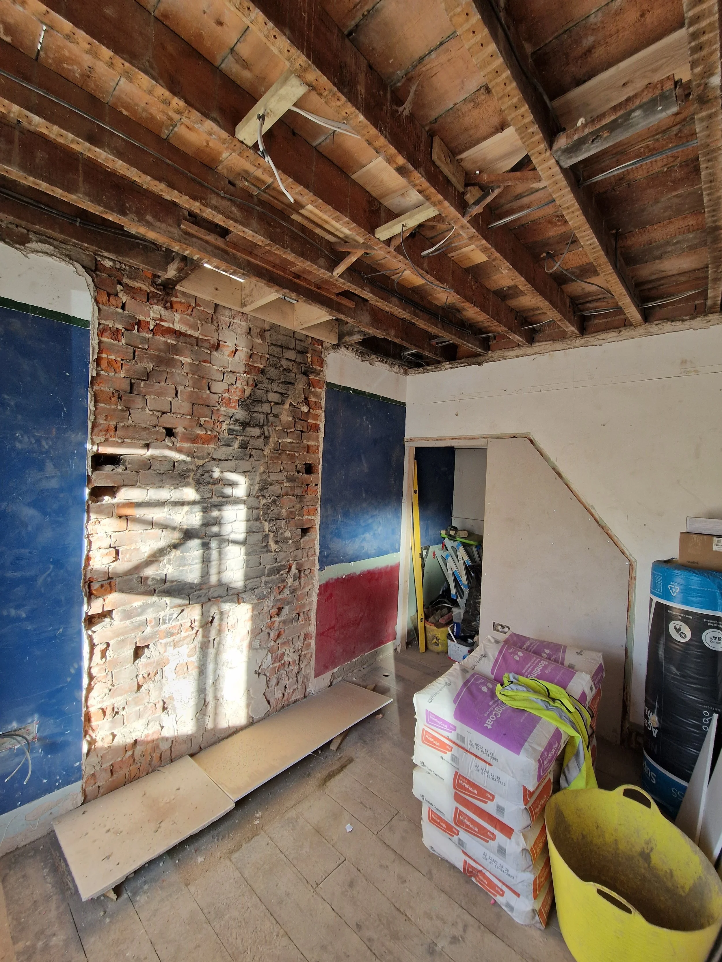 Interior of a room under renovation, with exposed brick wall, partially removed wall, construction materials, and tools.