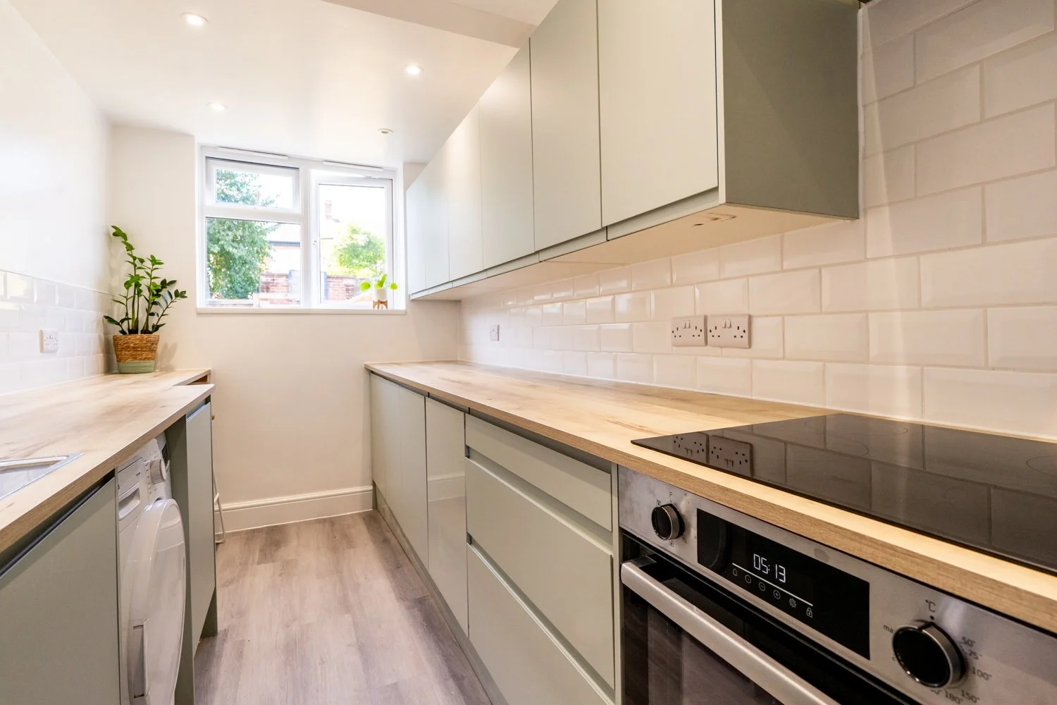 Modern kitchen with white cabinets, light wooden countertops, white subway tile backsplash, a window with a view of trees, and a potted plant on the left side.