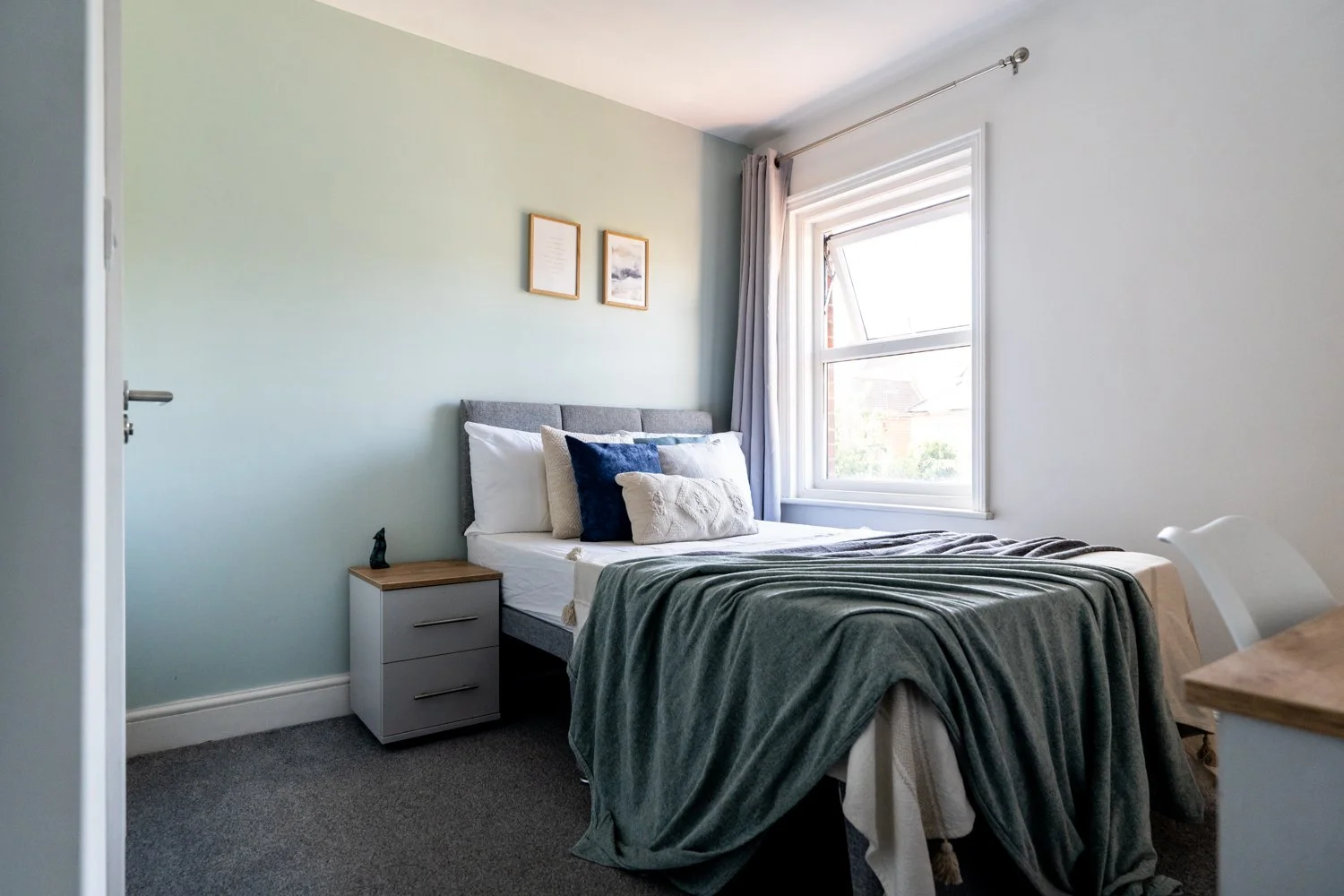 A bedroom with a neatly made bed with multiple pillows, a side table with a small decorative item, a window with curtains allowing sunlight in, and framed artwork on the mint green wall.
