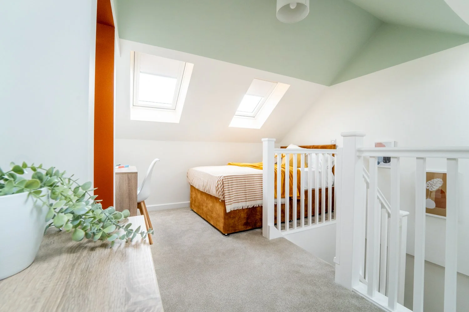 A cozy attic bedroom with two skylight windows, a bed with cream and brown striped bedding, a small wooden desk with a white chair, clothing on the bed, a white railing, and a potted plant on a wooden surface.