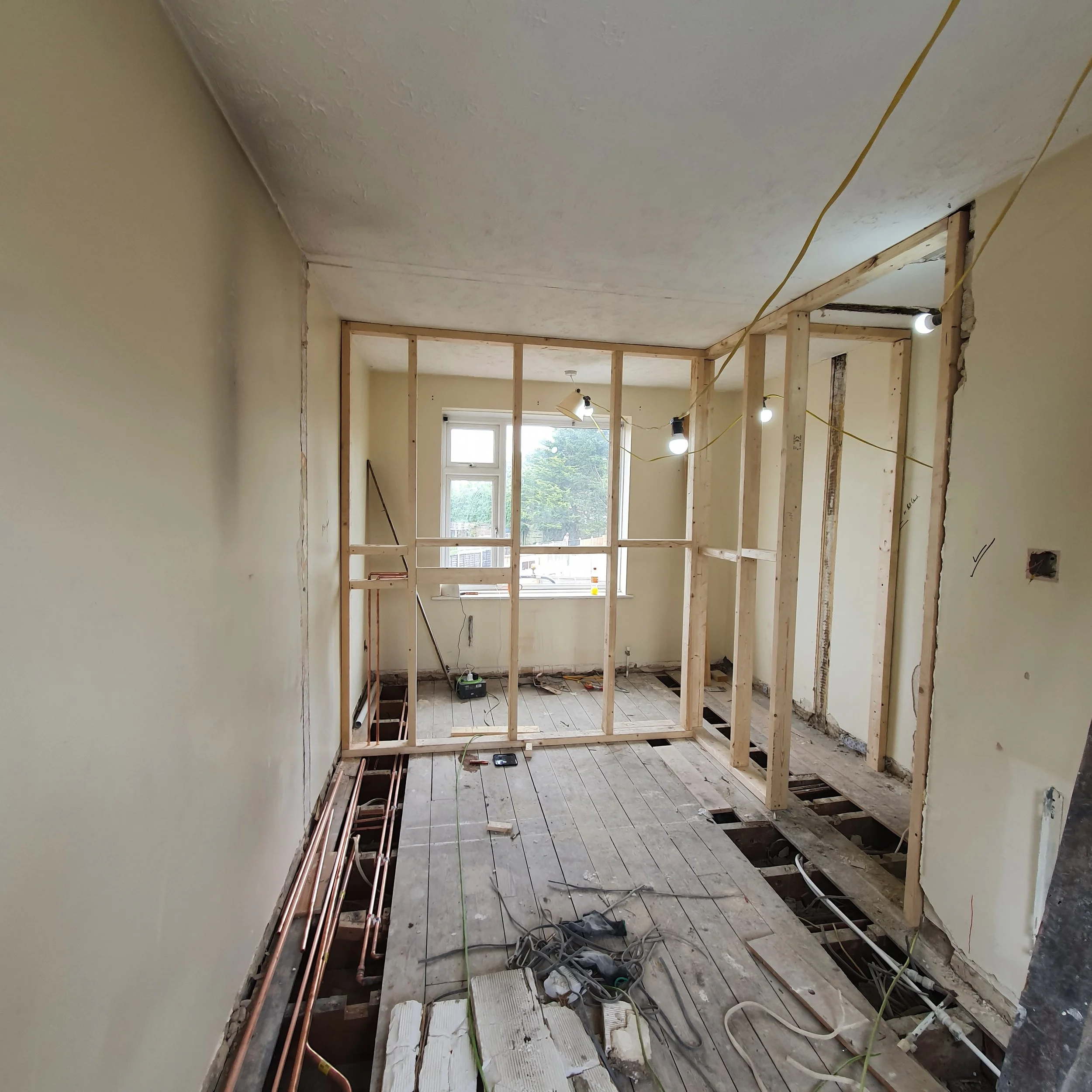 Interior space under construction with exposed wiring and heating pipes, wooden framing, and an unfinished floor, with light coming through a window.