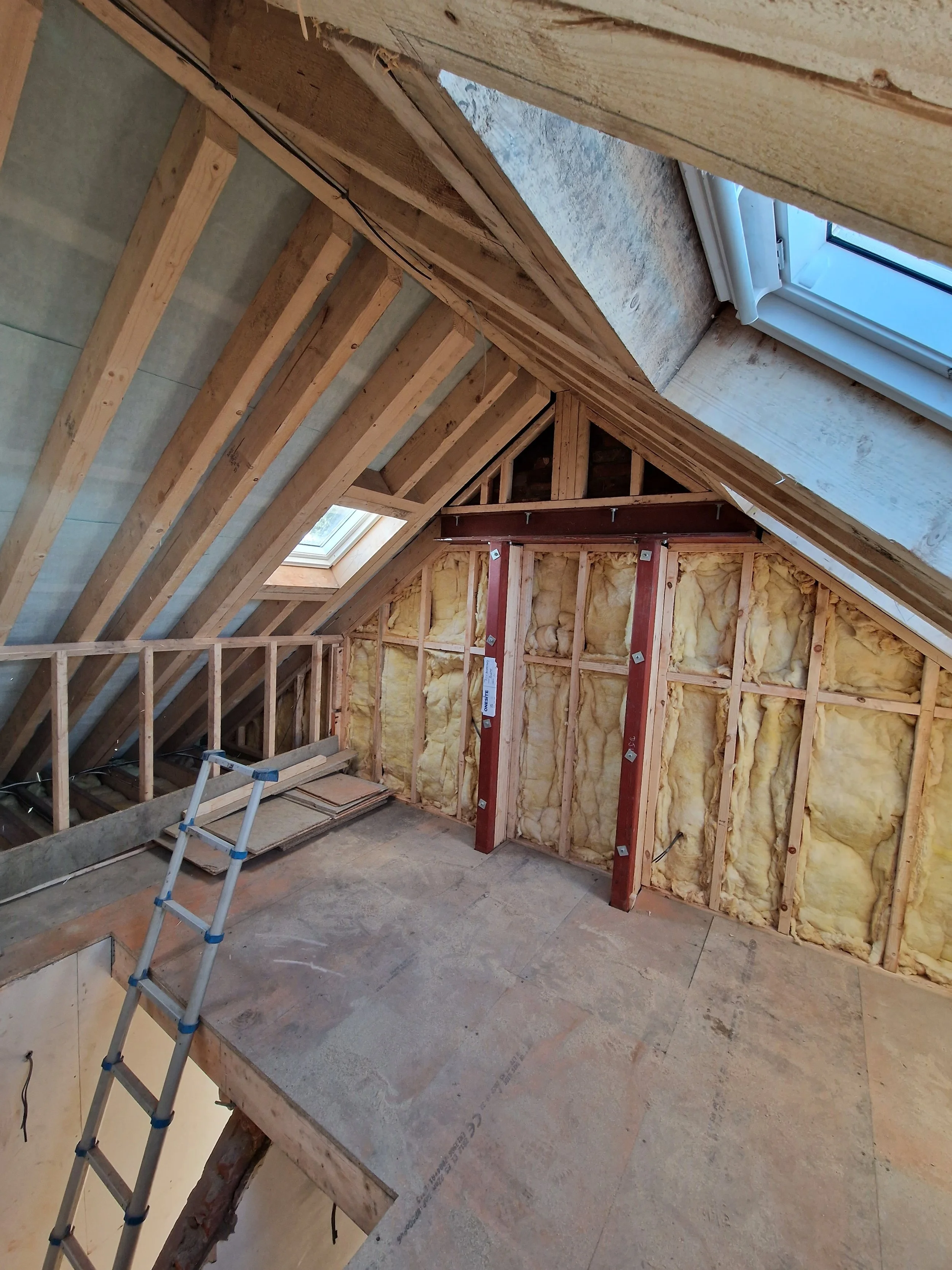 Attic space under construction showing exposed wooden framing, insulation, dormer window, and a small ladder.