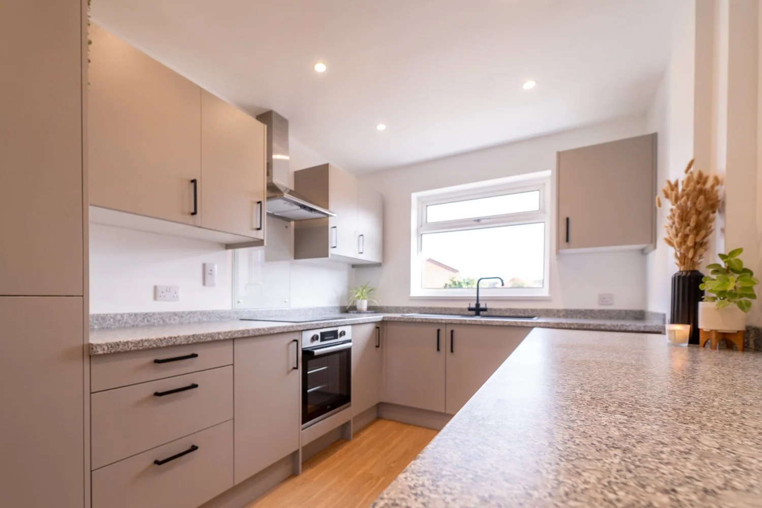 Modern kitchen with beige cabinets, granite countertops, black handles, an oven, oven hood, a window above the sink, and decorative plants on the counter.