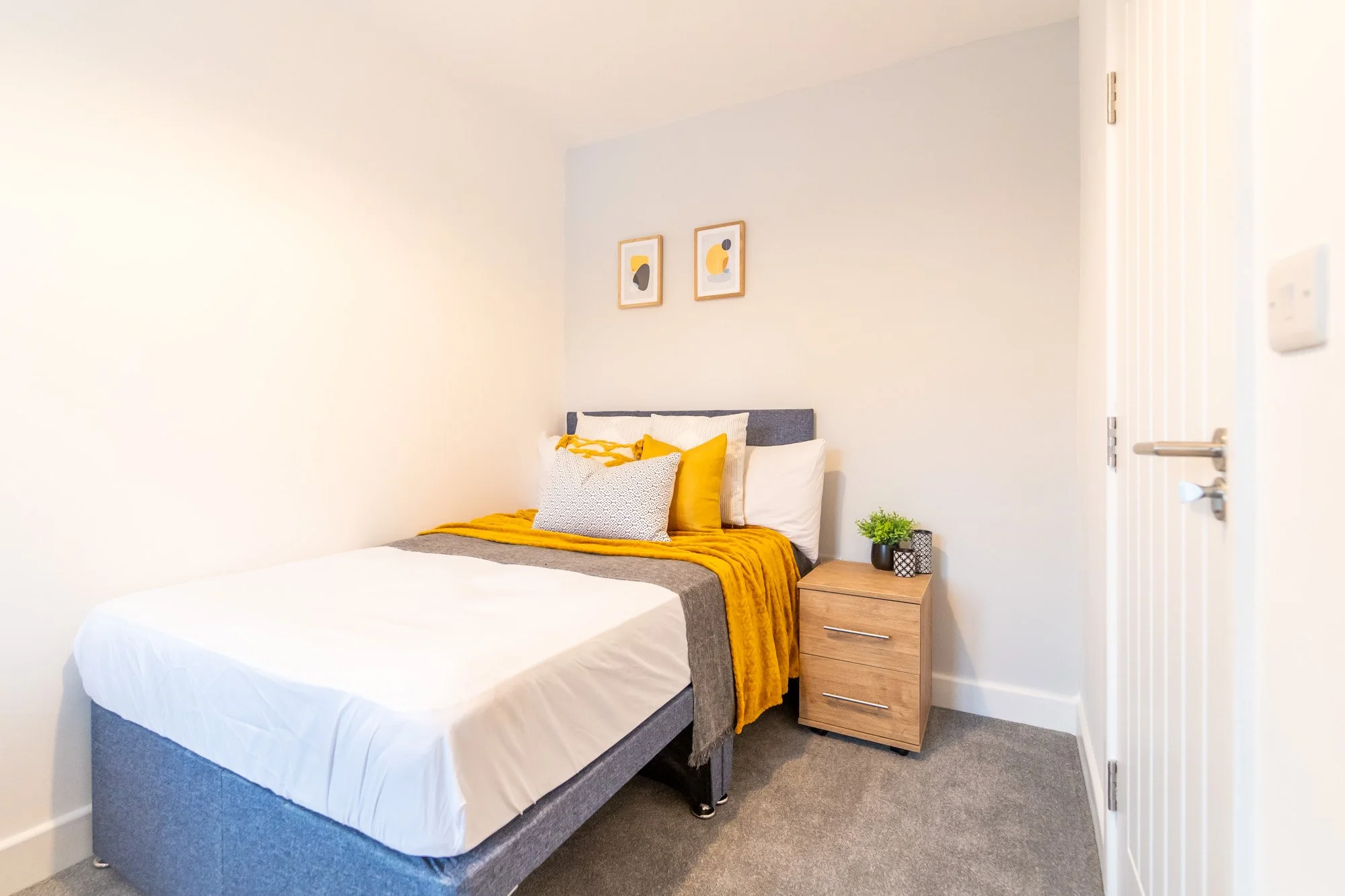 A neatly made bed with white and yellow pillows, a mustard yellow throw, and a gray headboard in a minimalist bedroom with a small wooden nightstand and two framed abstract art pieces on the wall.