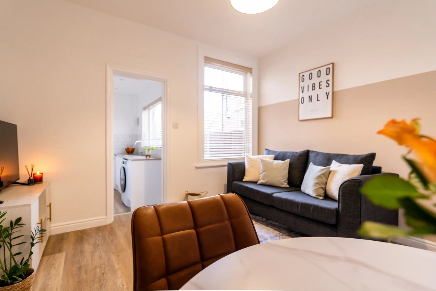 Living room with black sofa, white pillows, wall art reading 'Good Vibes Only,' window with blinds, and dining table with brown leather chair.