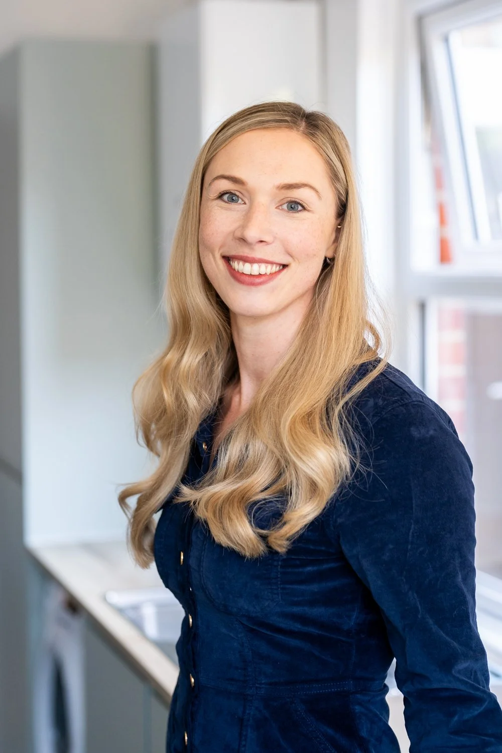 A woman with long blonde hair, blue eyes, and a bright smile standing indoors near a window.