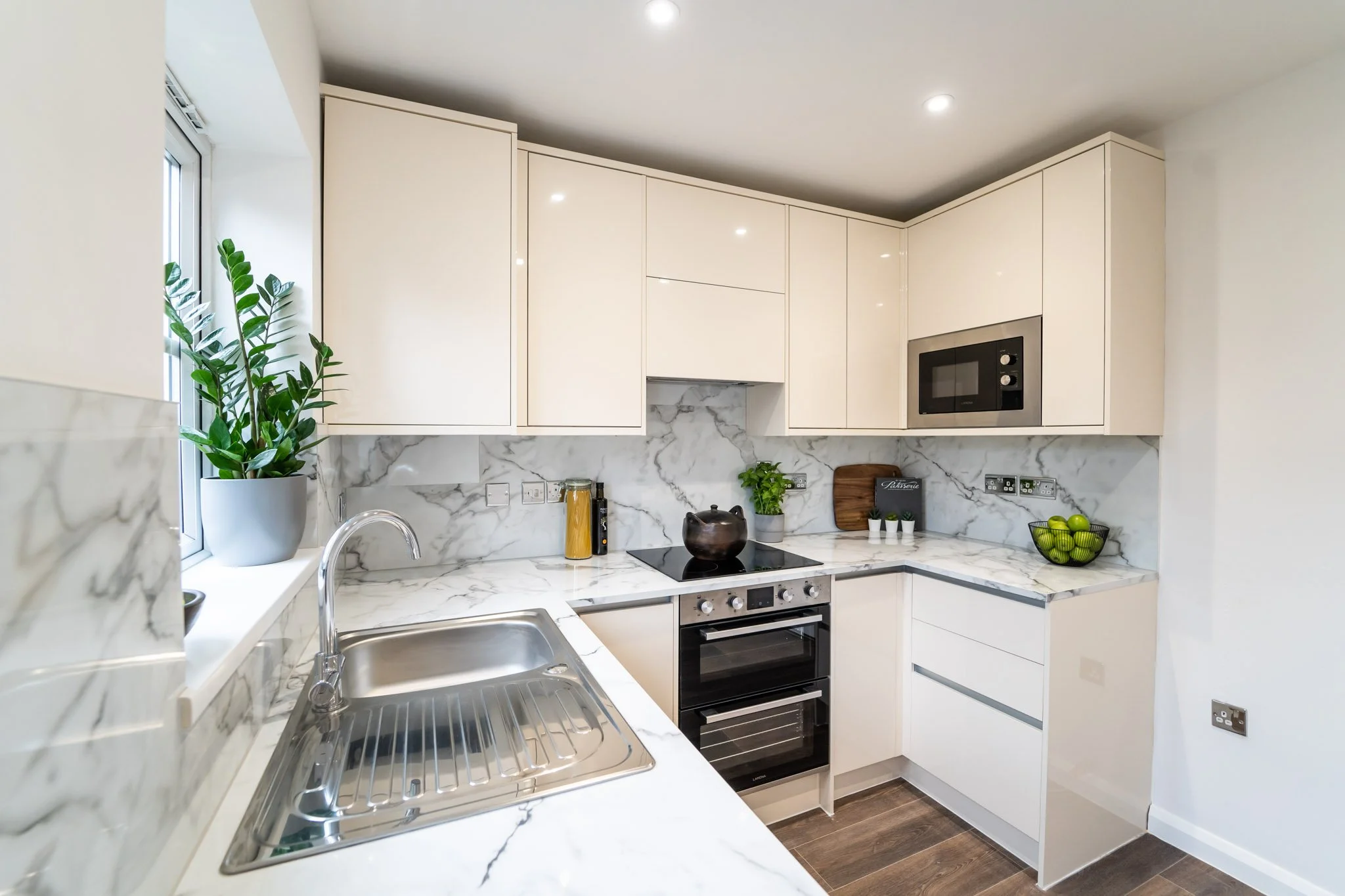 Modern kitchen with white cabinets, marble countertops, and a stainless steel sink near a window with green plants, black appliances, and decorative items.
