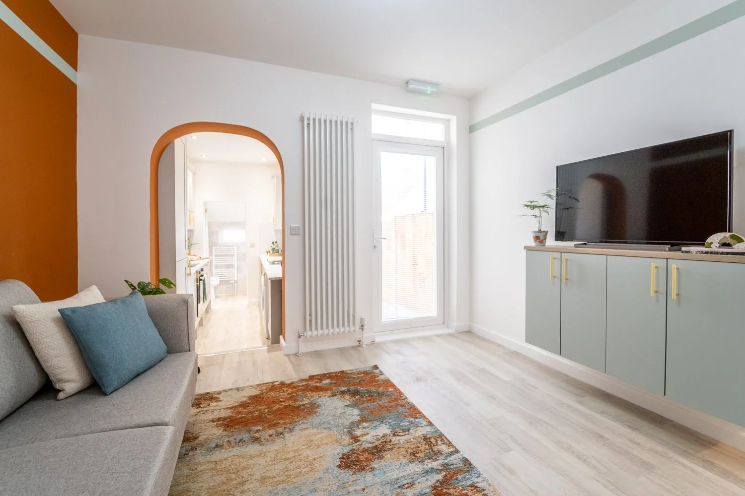 Living room with a gray sofa, pillows, a colorful area rug, a wall-mounted TV, and a door leading to a bright room with a kitchen area in the background.