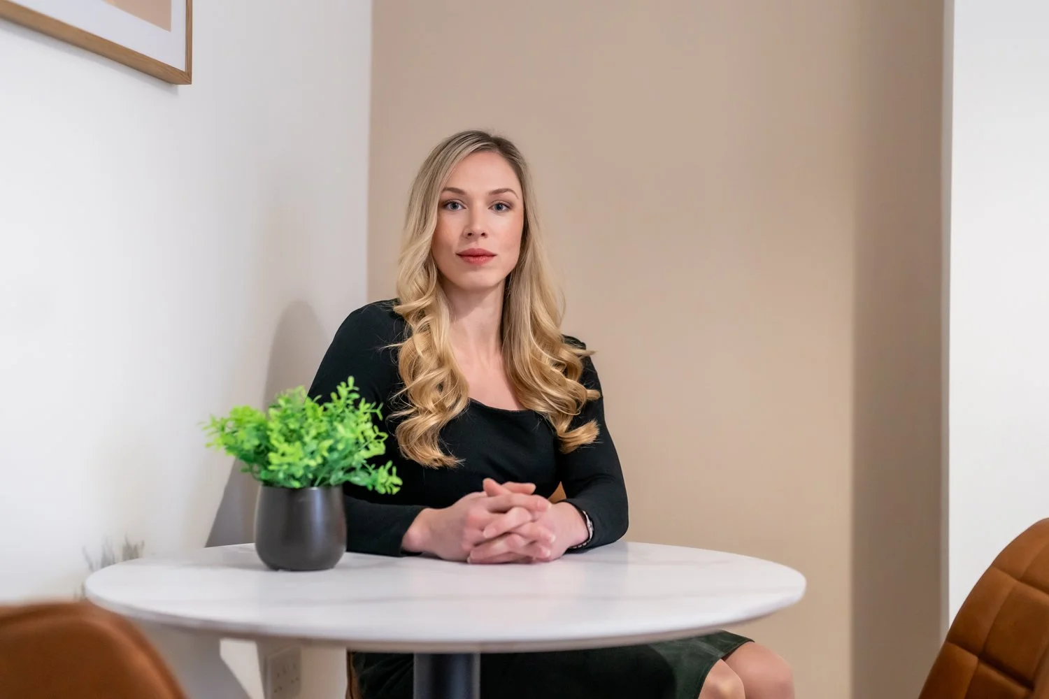 A woman with long blonde hair, wearing a black top, sitting at a white round table with a small green potted plant, in a minimalistic room with beige and white walls.