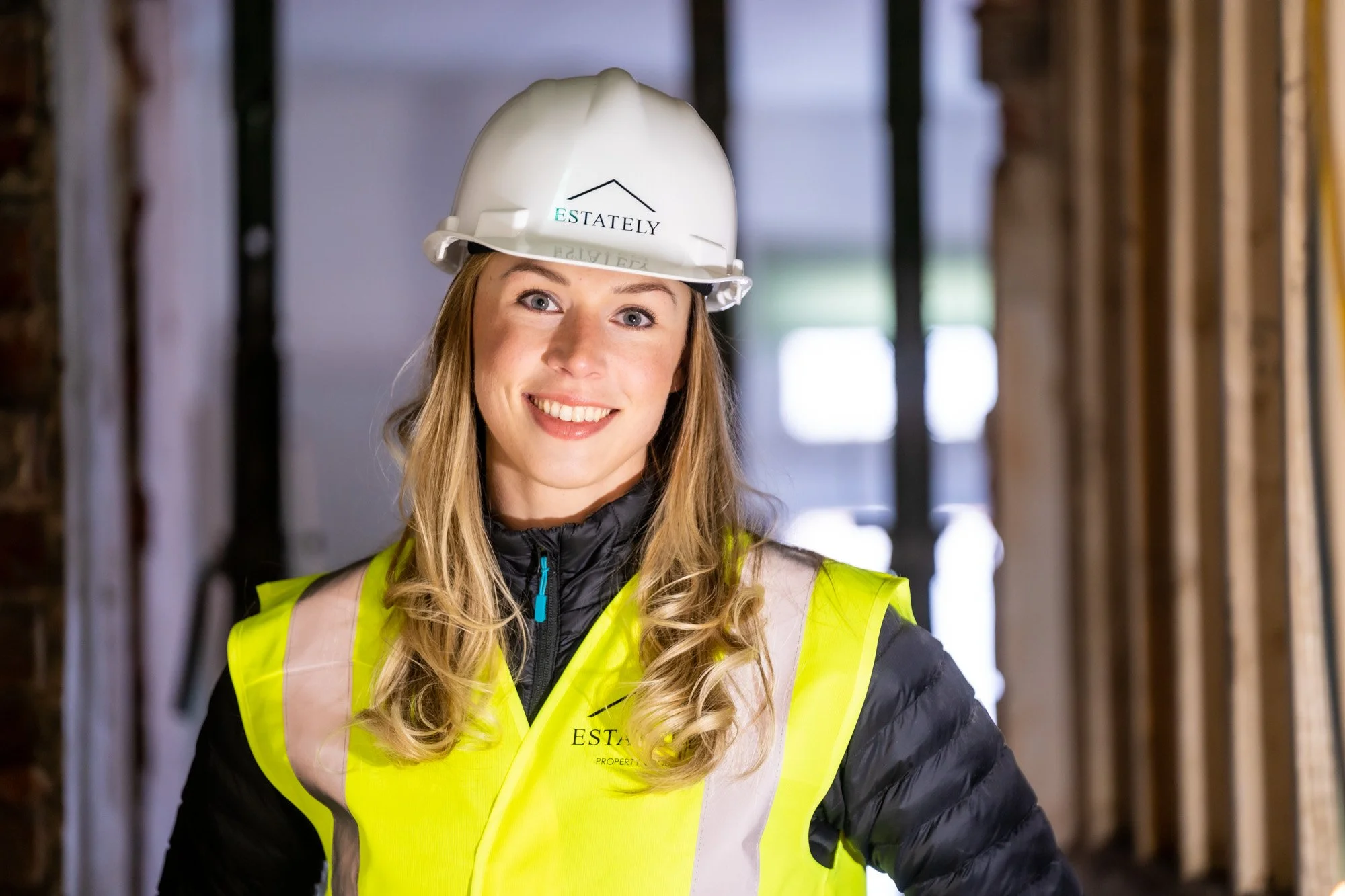 A smiling woman wearing a white hard hat and yellow safety vest inside a construction site with wooden framing.