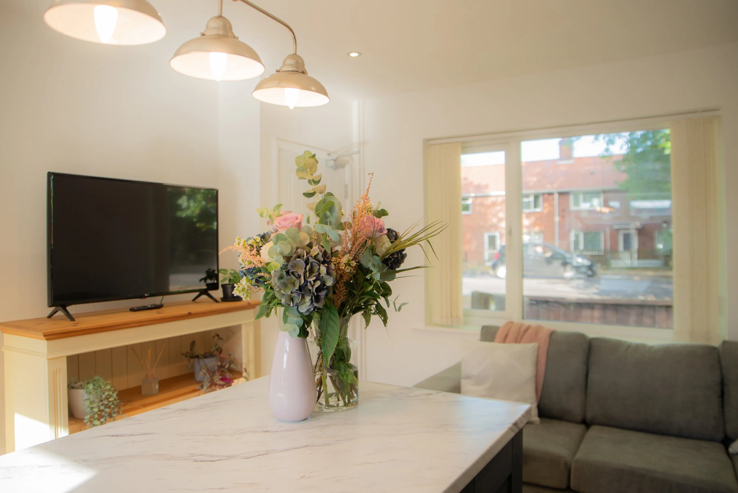 Living room with a bouquet of flowers on a white marble table, a television on a wooden stand, a gray sectional sofa with pink and white pillows, and a large window showing a street view outside.