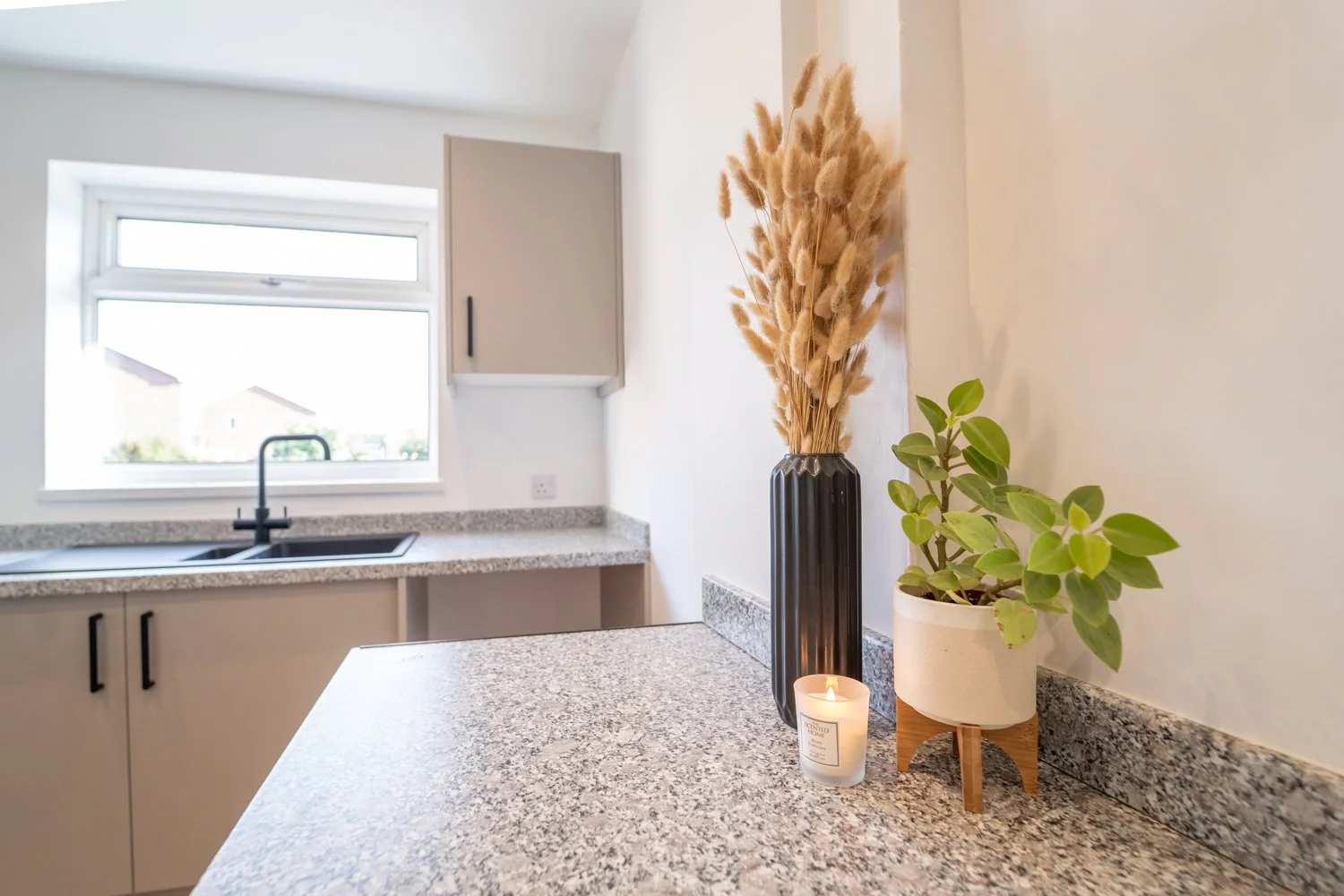 Decorative arrangement on a kitchen counter featuring a tall black vase with dried pampas grass, a potted green plant, and a lit white candle in a glass container, with a window, beige cabinets, and granite countertops in the background.