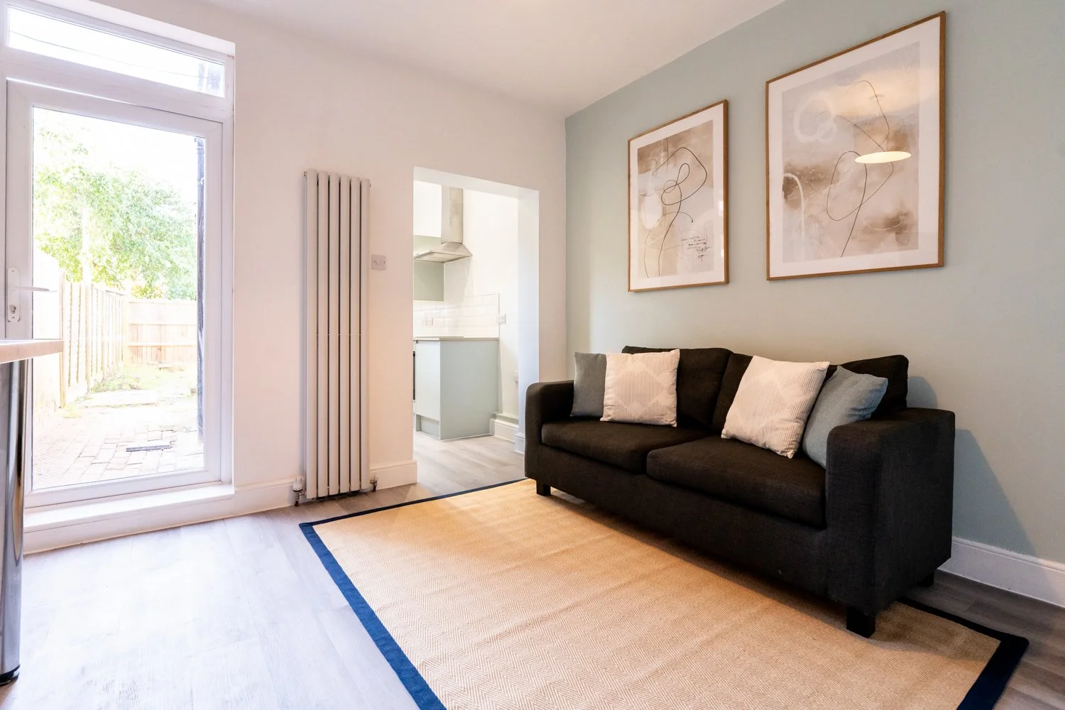 Living room with a black sofa, three cushions, wall art, a beige rug with a blue border, a glass door leading outside, and a view into the kitchen.