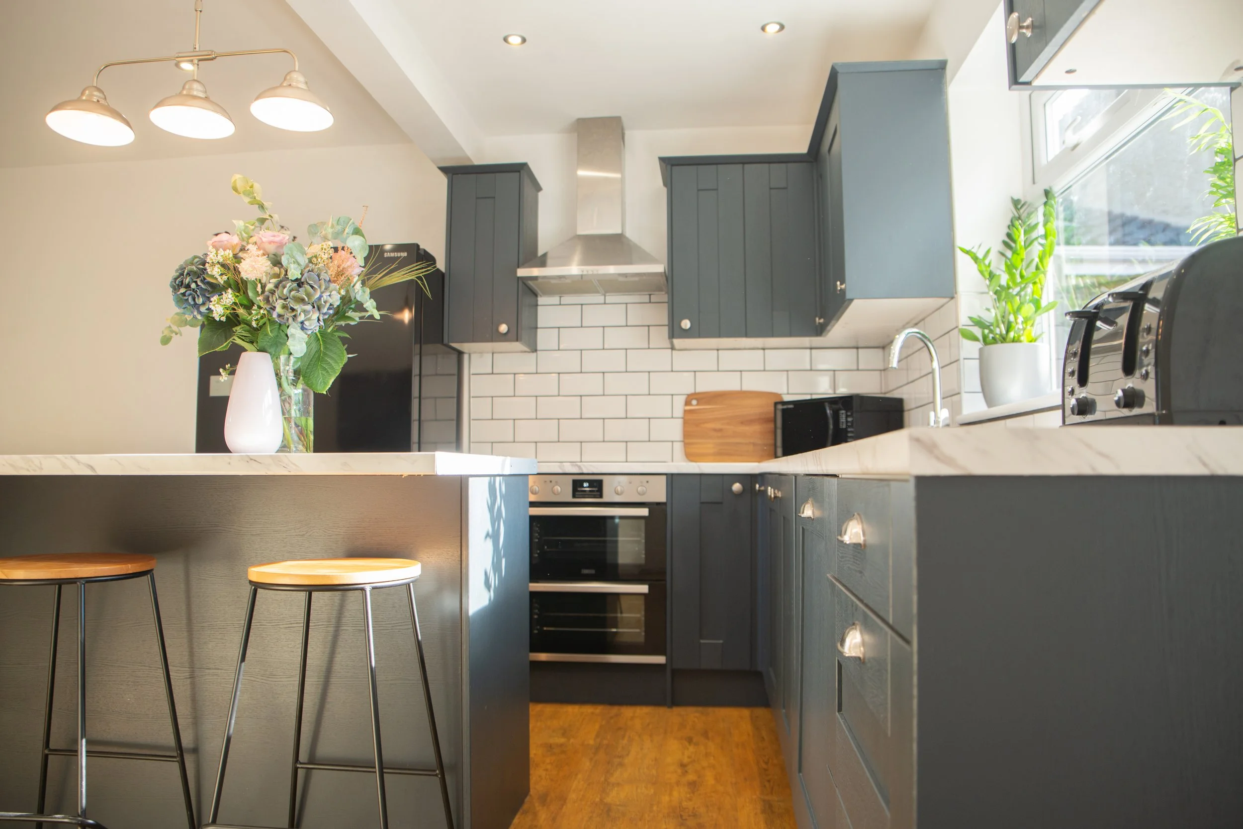 Modern kitchen with dark gray cabinets, white subway tile backsplash, and wooden accents. Features include a kitchen island with two stools, a floral vase, potted plants, and various appliances like an oven, microwave, and toaster.
