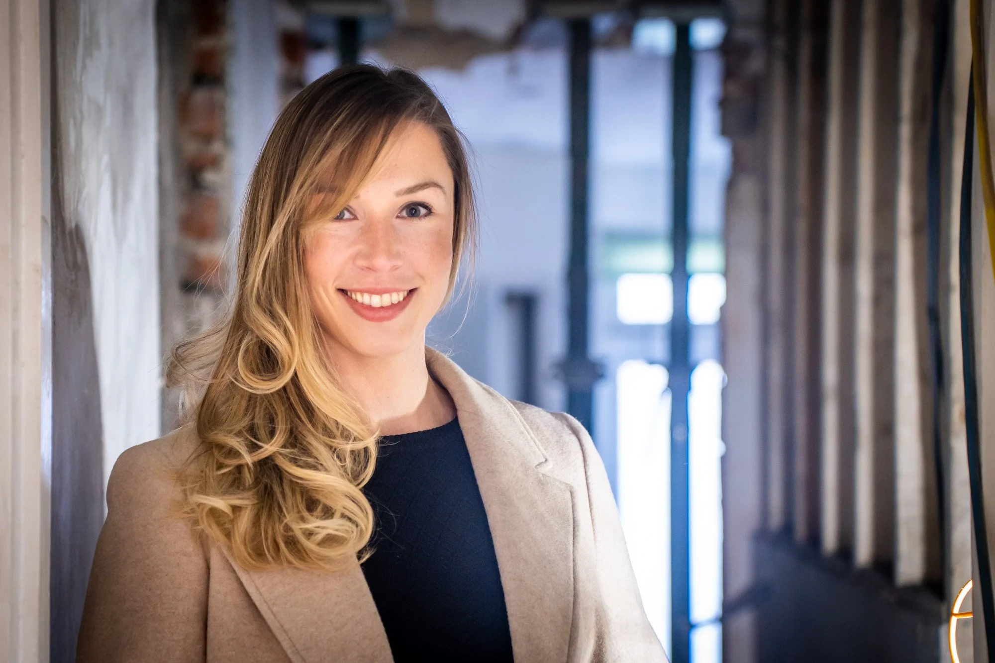 A smiling woman with long blonde curly hair, wearing a beige coat and black top, standing in a rustic hallway with brick and wooden walls.