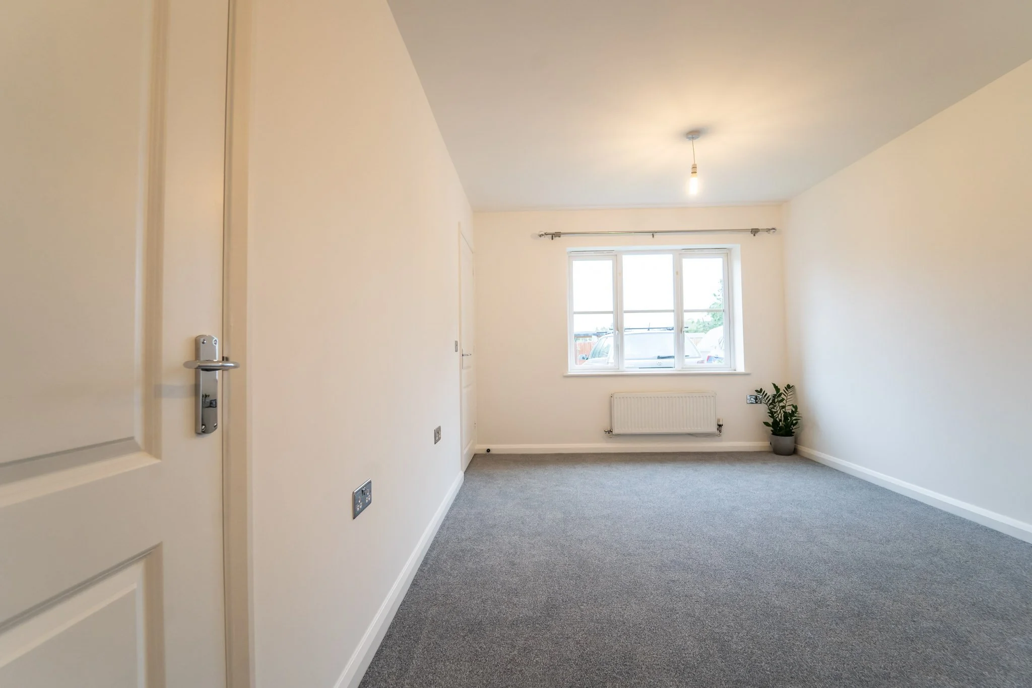 Empty room with light-colored walls, gray carpet, a window with a view outside, a radiator, a small green potted plant, and a ceiling light with a bulb.