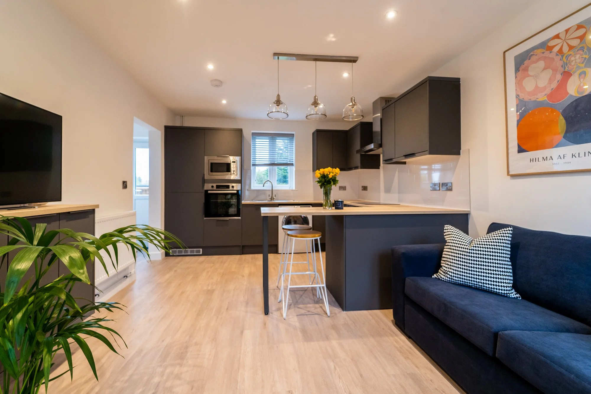 Modern open-concept living room and kitchen with black cabinetry, wooden flooring, a black sofa with a checkered pillow, a TV on a stand, and a dining table with stools, decorated with a bouquet of yellow flowers.