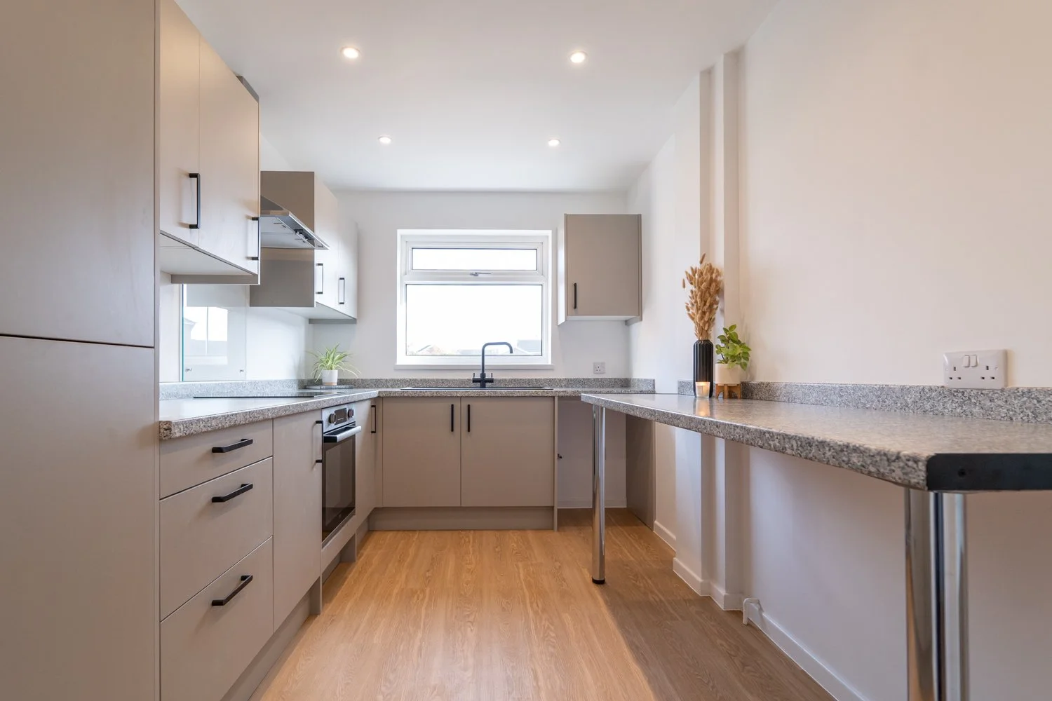 Modern kitchen with beige cabinets, gray granite countertops, a window above the sink, wooden floor, and decorative plants.