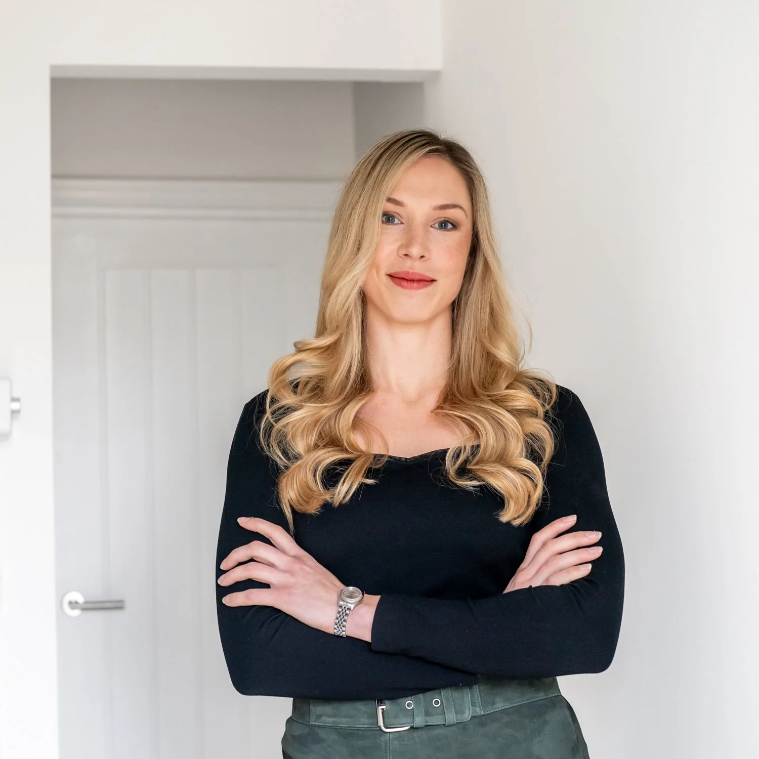 A woman with long blonde hair and light makeup standing with arms crossed against a white wall, wearing a black long-sleeve top and a silver watch.