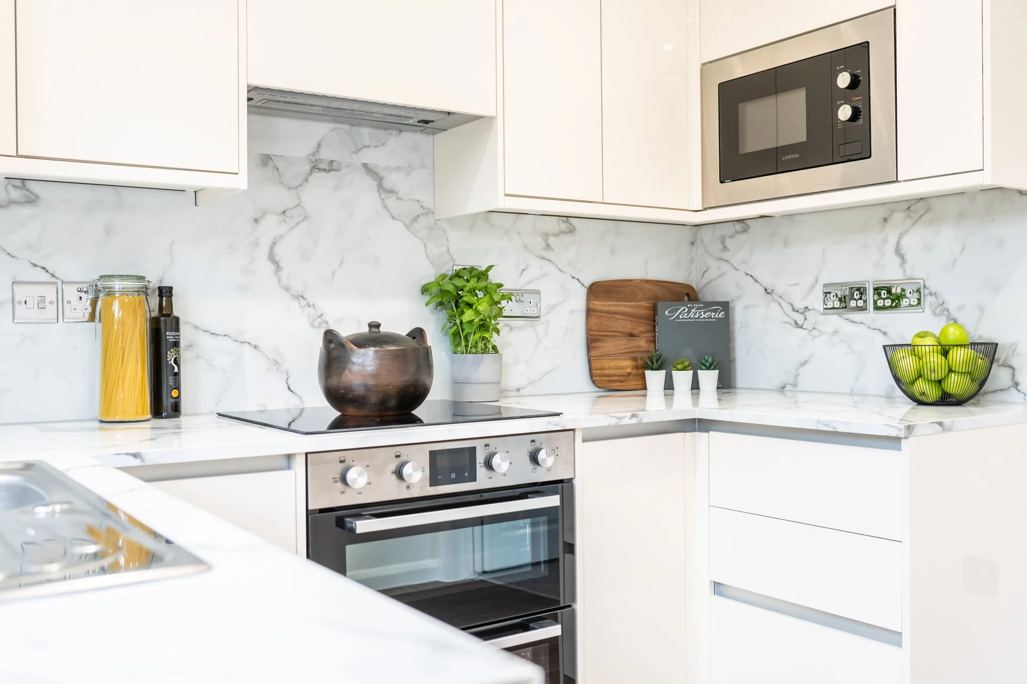 Modern kitchen with white marble countertops and backsplash, stainless steel oven, microwave, and black cooking pot on stove, with potted herbs, green apples in a wire basket, and a cutting board with a small book and decorative plants.