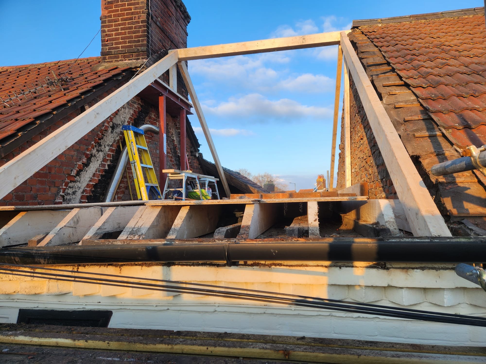 Construction work on roof with wooden framing and ladders, brick chimneys, and blue sky in the background.