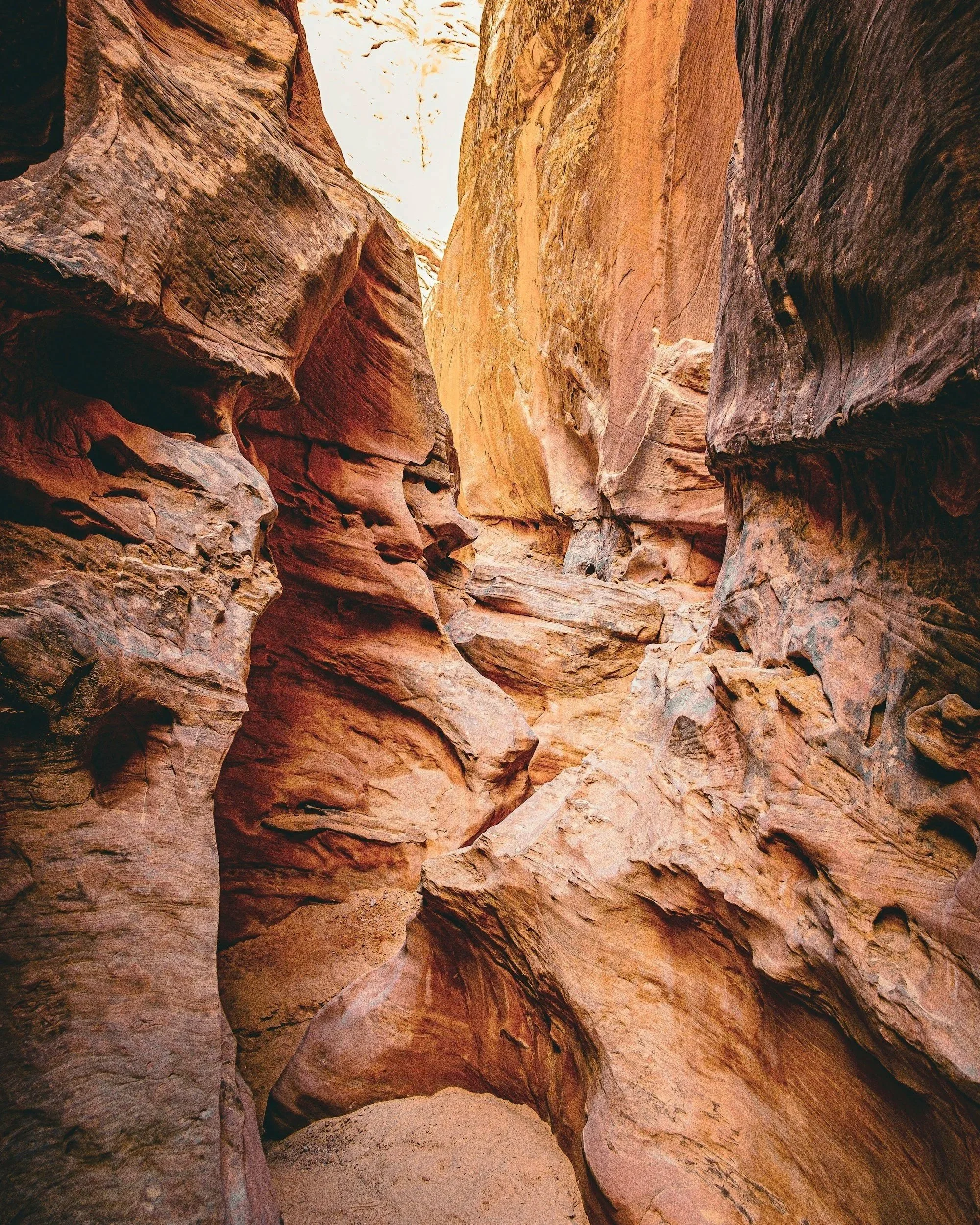 A narrow, winding slot canyon with smooth, layered sandstone walls in shades of red, orange, and tan. Sunlight filters down from above, illuminating parts of the canyon and casting shadows on the textured surfaces.