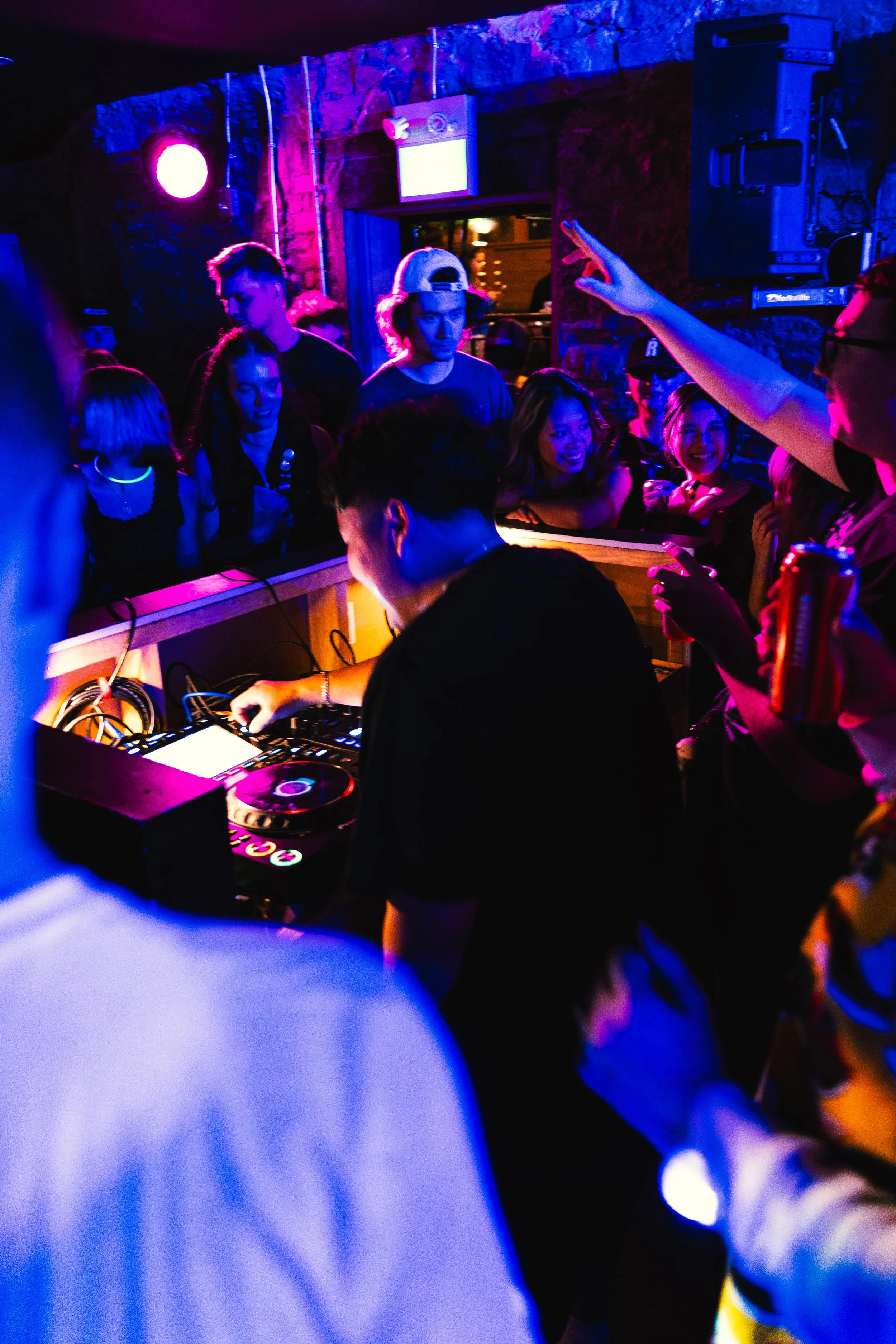A DJ performs at a nightclub with colorful purple, pink, and blue lighting. The crowd is dancing and enjoying the music, with some people holding drinks and taking photos.