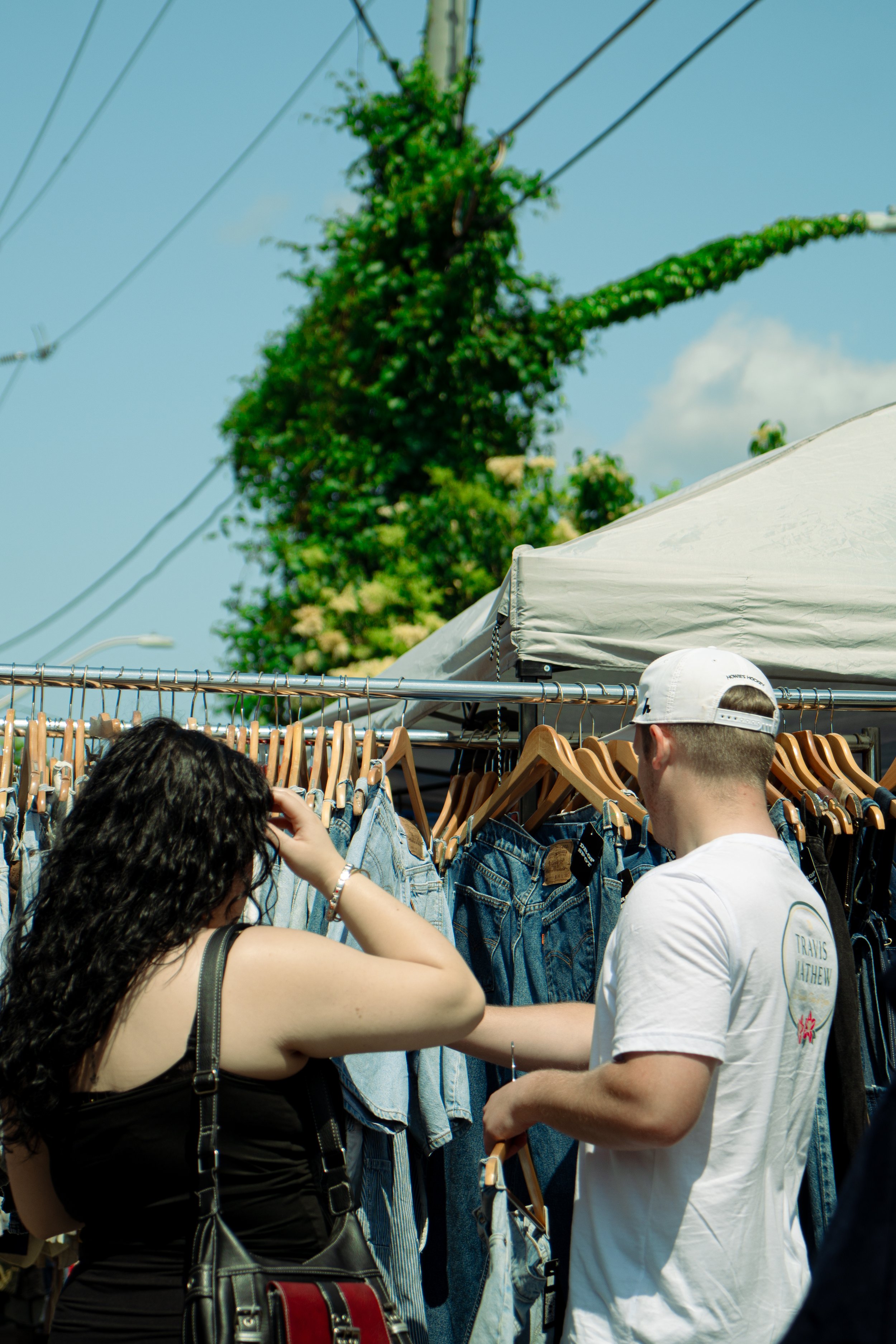 A young woman and a young man browsing through clothes at an outdoor market stall with hanging denim jeans and shorts. The woman has curly dark hair, wears a black top, and carries a black purse. The man has light skin, wears a white cap backward and a white t-shirt.