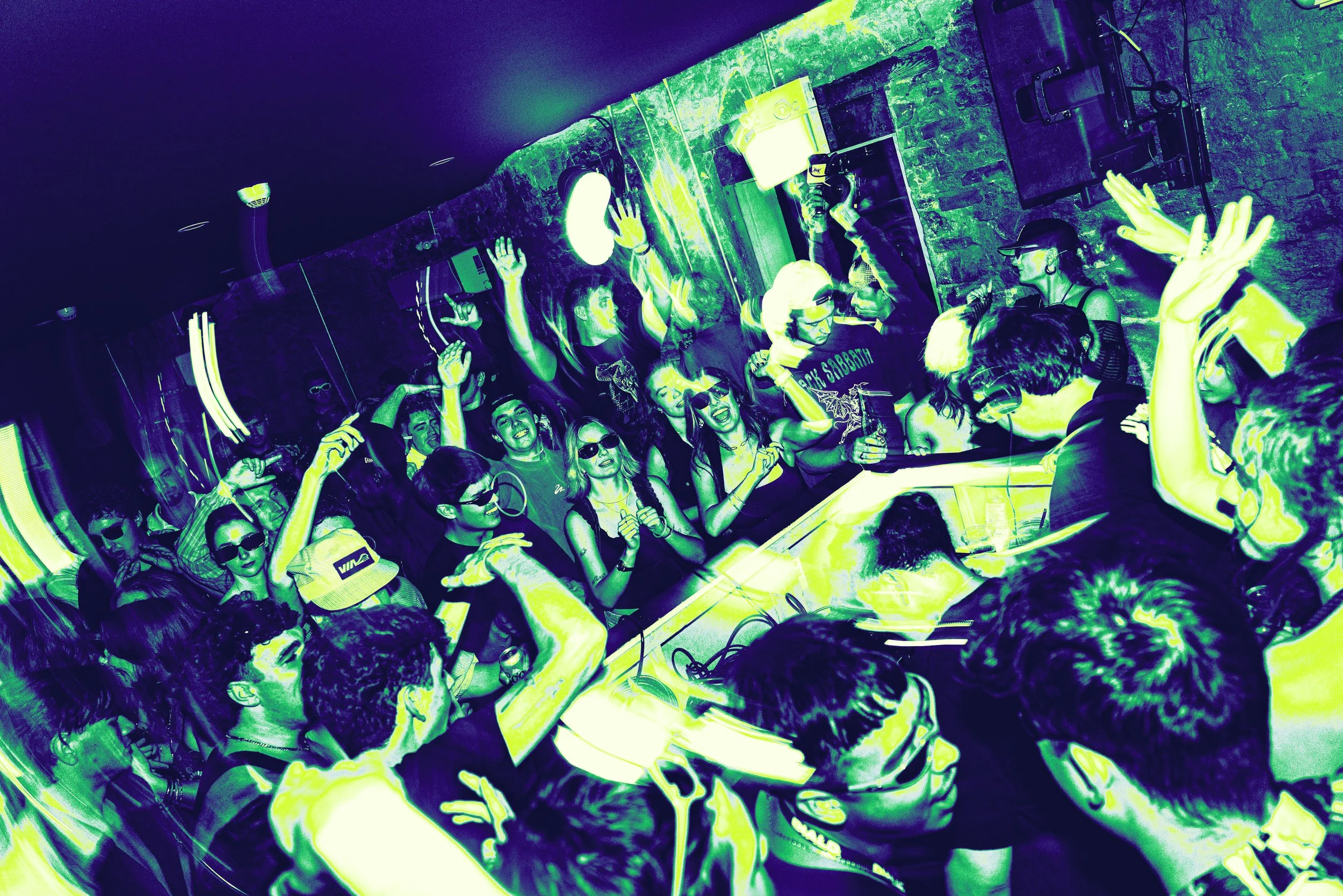 A lively party scene with many young people dancing, singing, and enjoying music at a nightclub. Some individuals are wearing sunglasses, raising their hands, or holding drinks, with a DJ playing music in the foreground. The room features wall art an