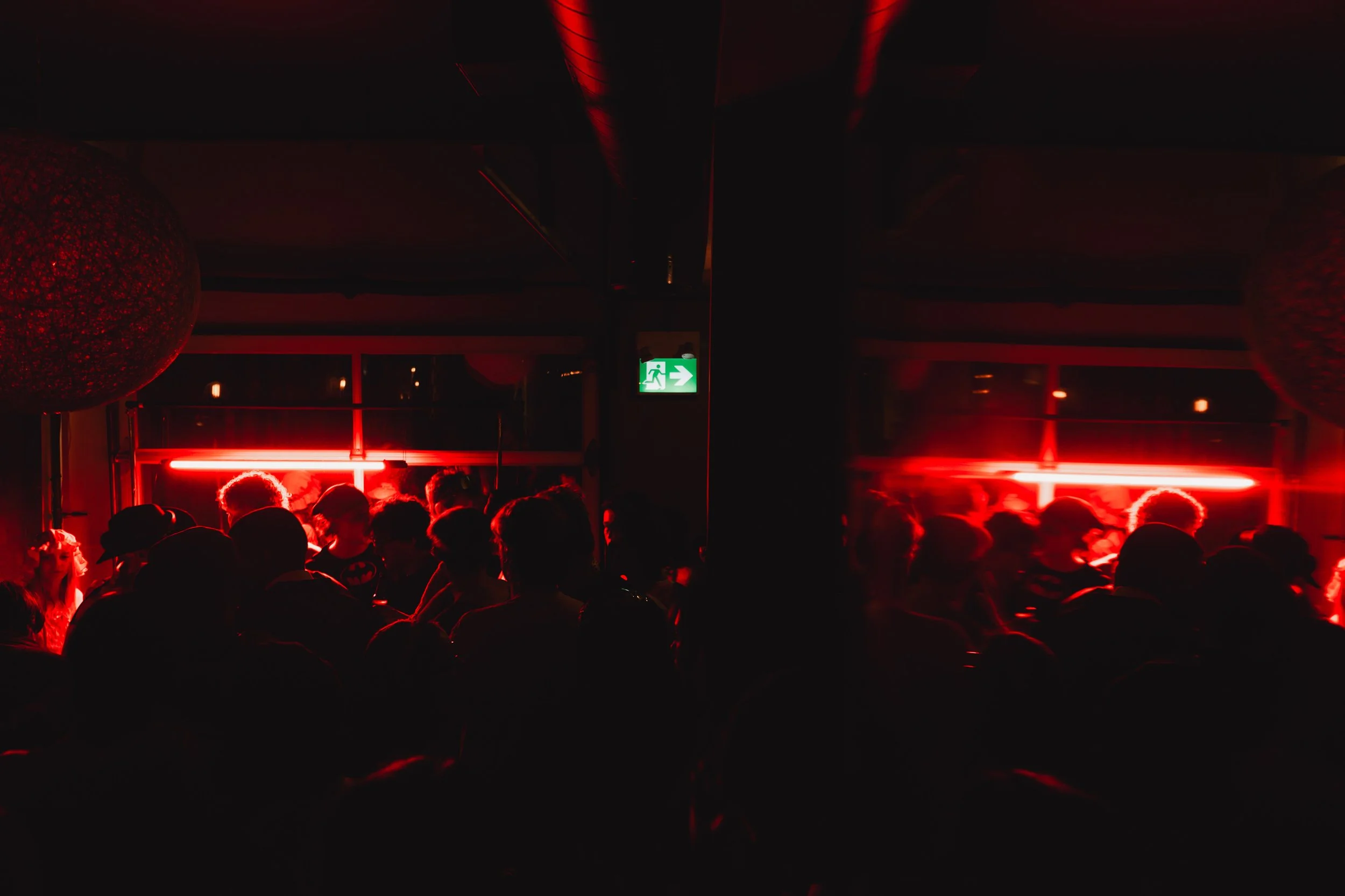 Dark indoor party scene with people in silhouette, illuminated by red neon lights, with an exit sign visible at the back.