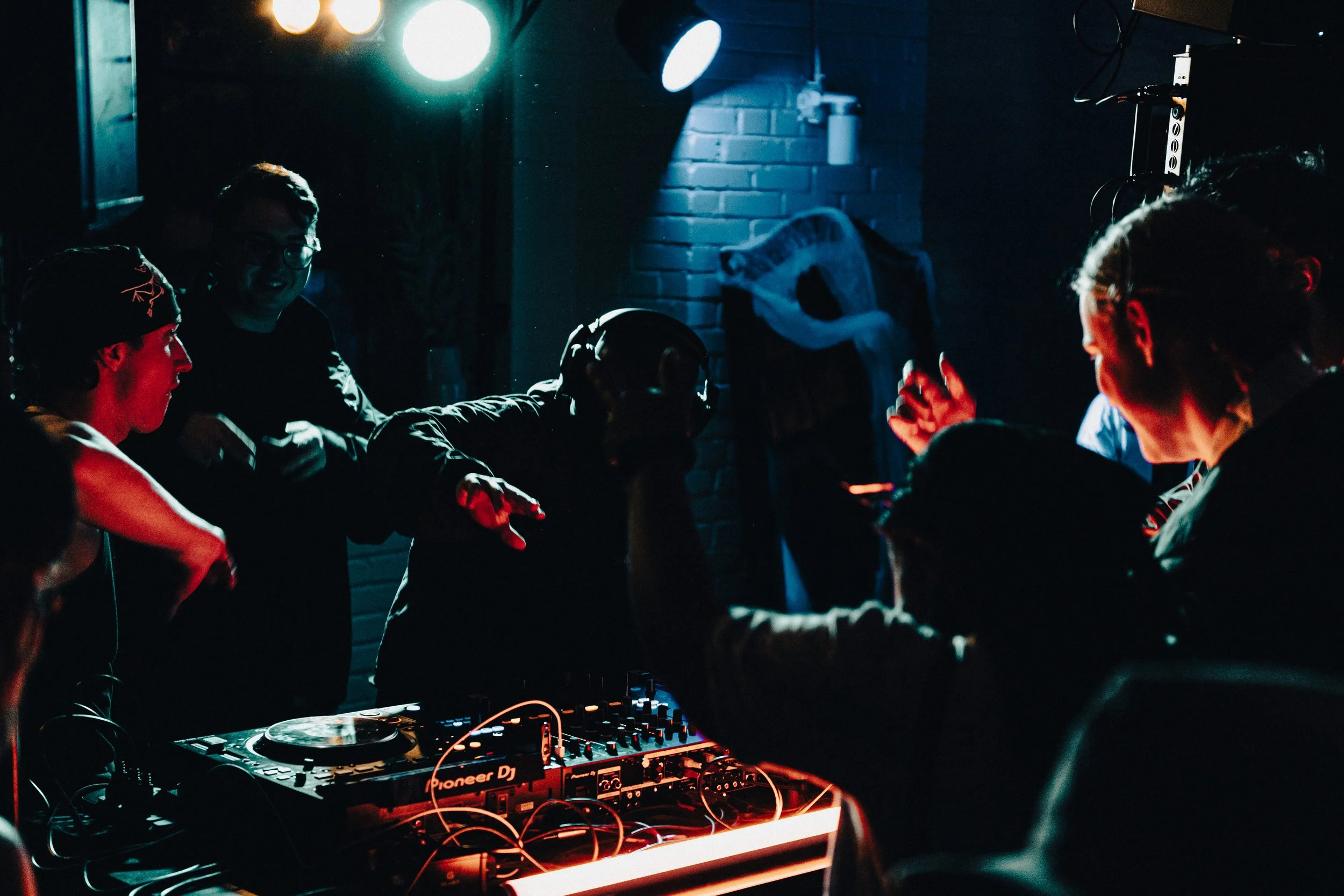 People dancing and DJing at a nightclub with colorful lighting and a brick wall in the background.
