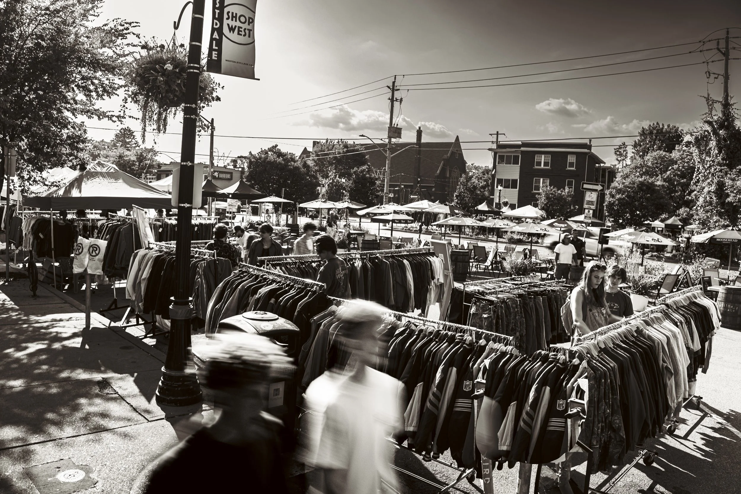 Black and white photo of an outdoor street market with clothing racks, umbrellas, and shoppers walking among the stalls.
