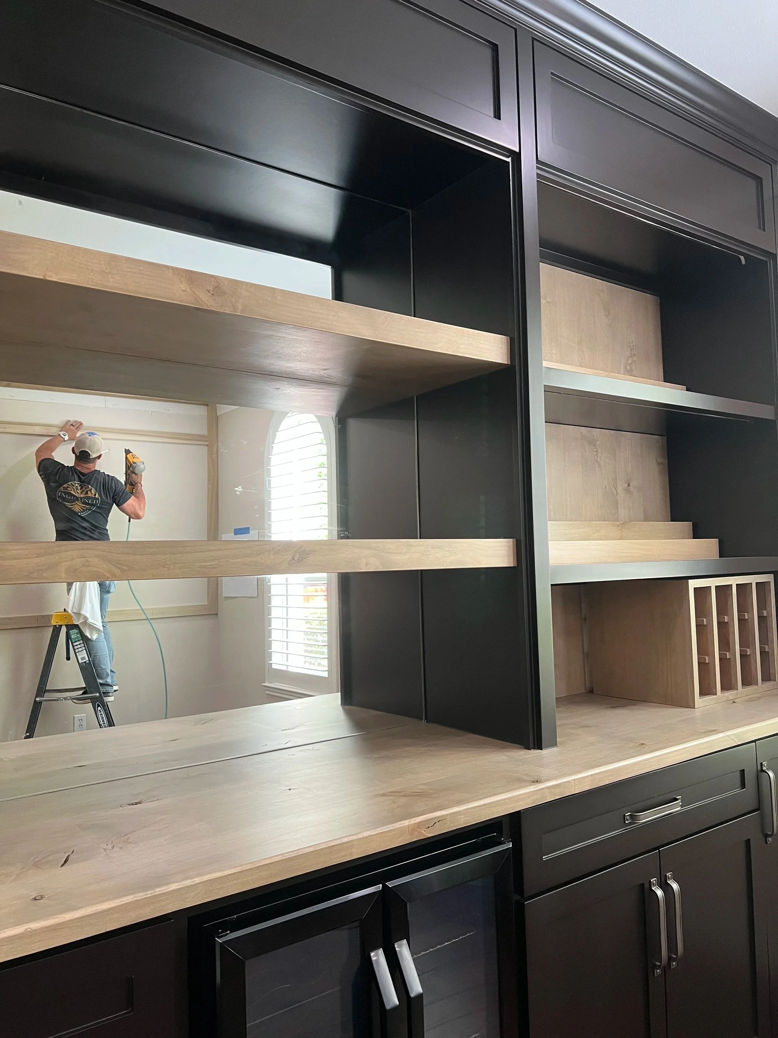 A person working on a construction project is seen through a cut-out in a black-and-wood kitchen cabinet. The person is standing on a ladder, using a drill, and is wearing a black shirt and a white cap. The room has a window with white shutters and light-colored walls.