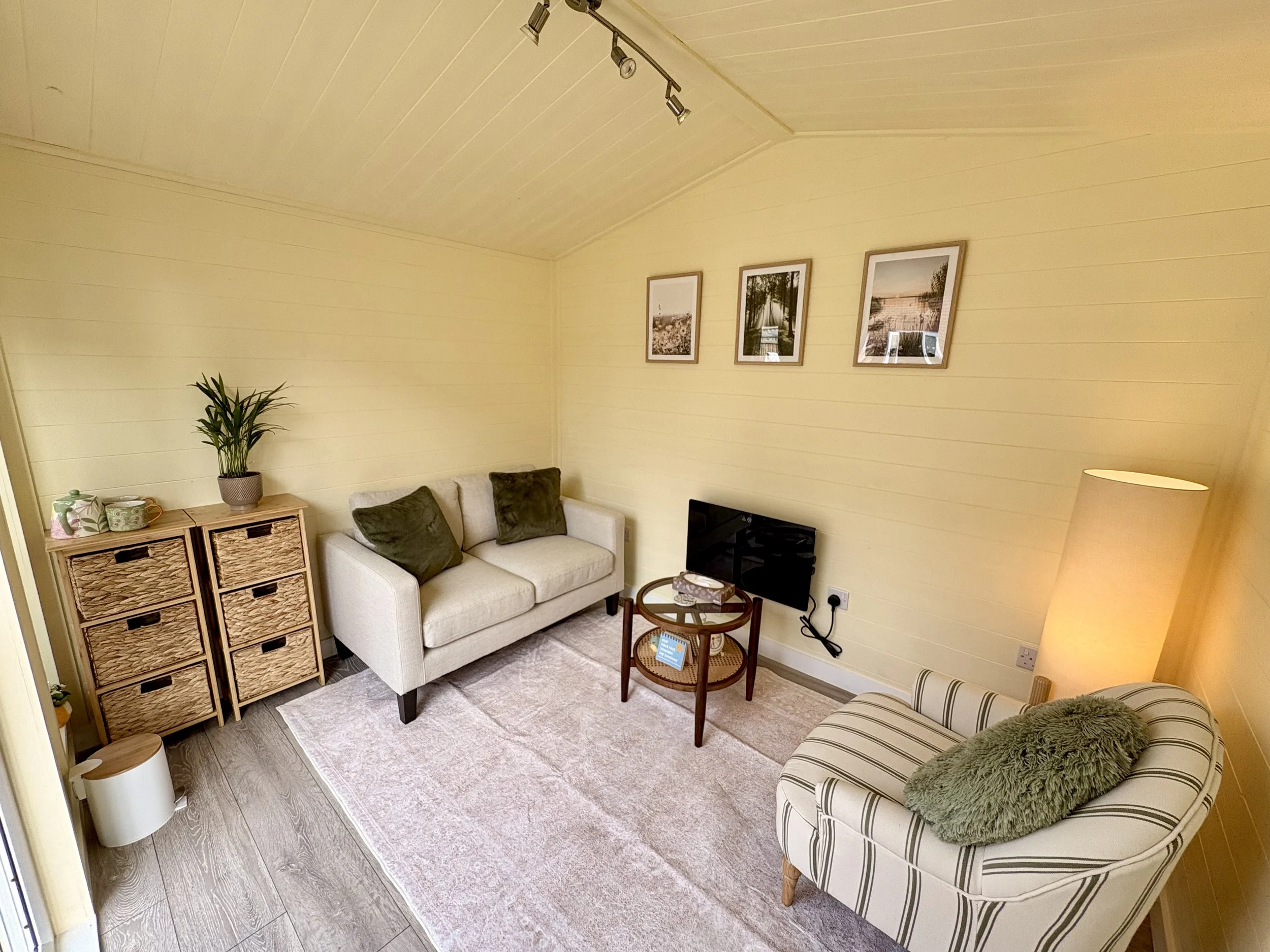 A cosy, private therapy space with cream wooden panelled walls, a cream soft sofa and green striped arm chair, both with green pillows. A tall soft glowing light, three picture frames of nature in the summer, and wicker drawers with plant and teapot.