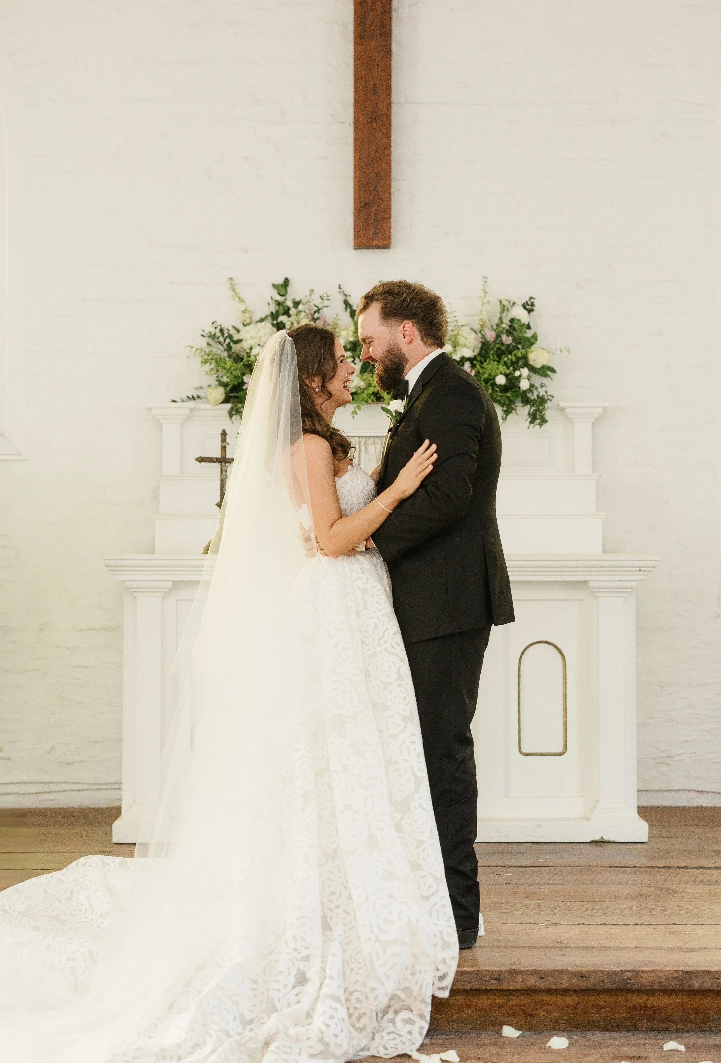 A bride and groom standing close together, smiling, and touching foreheads during their wedding ceremony in a white chapel with floral arrangements and a wooden cross in the background.