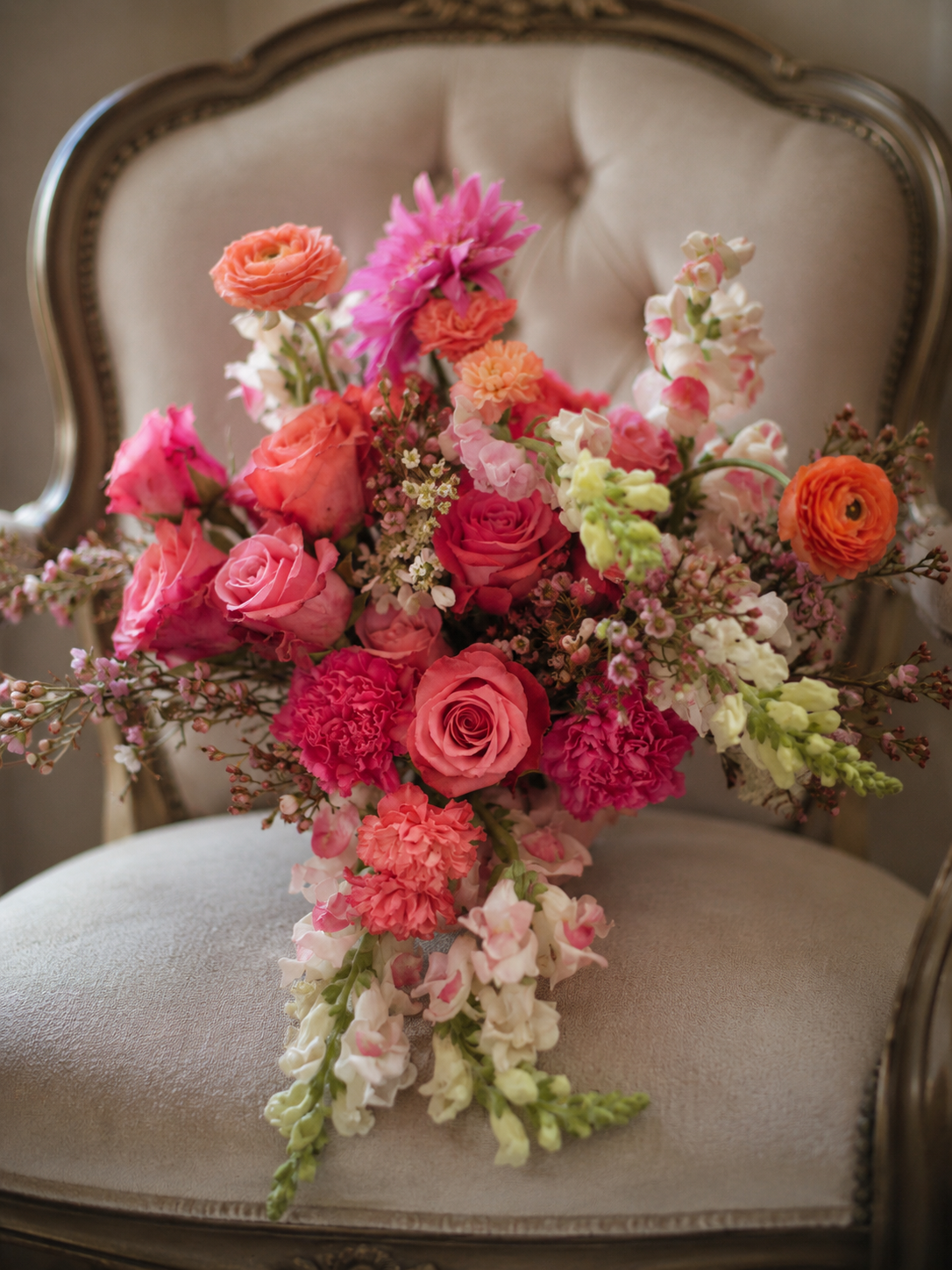 A bouquet of pink, orange, and white flowers resting on a vintage-style upholstered armchair with carved wooden trim.