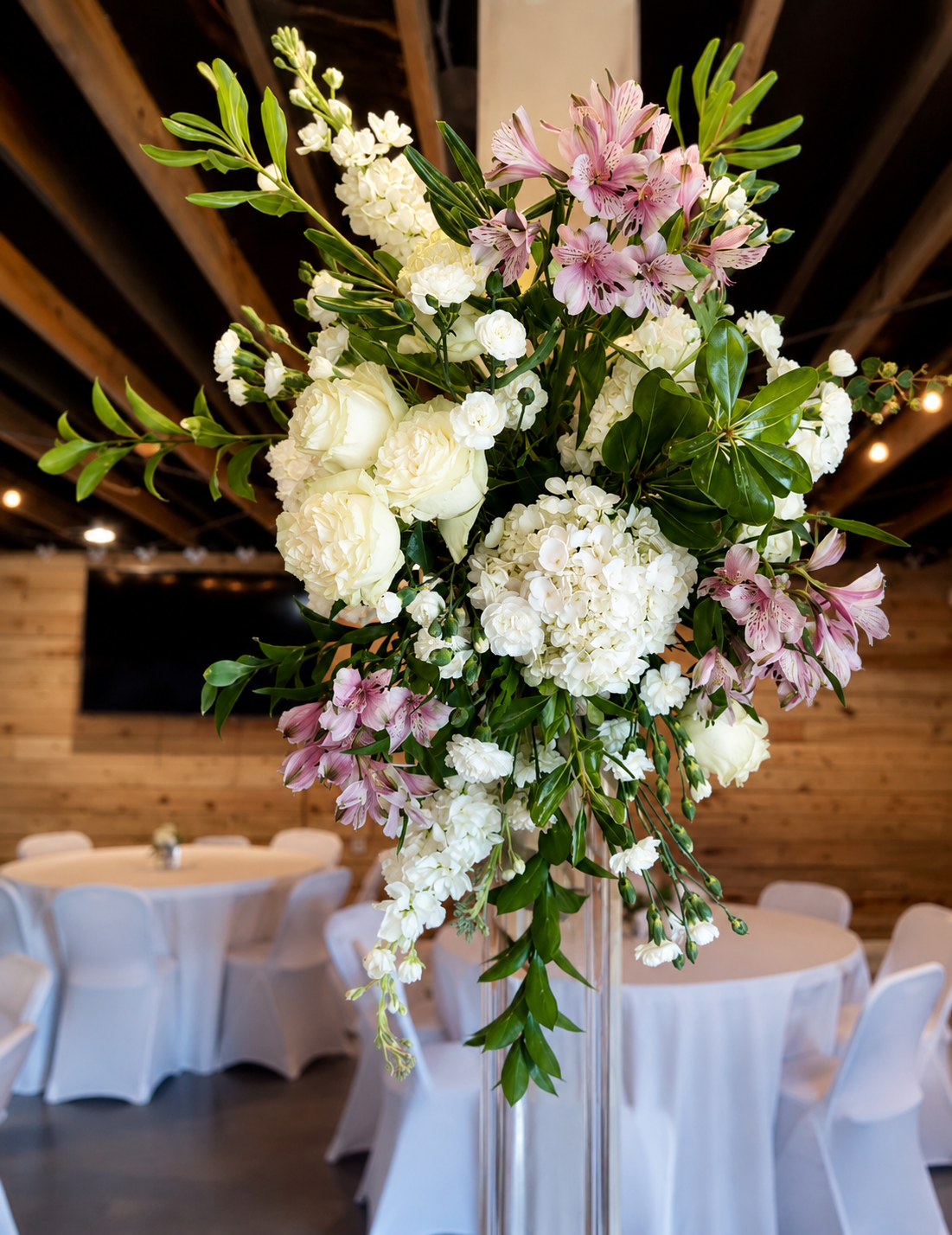A tall floral centerpiece with white and pink flowers and green leaves on a table in a decorated venue with wooden walls and ceiling beams.