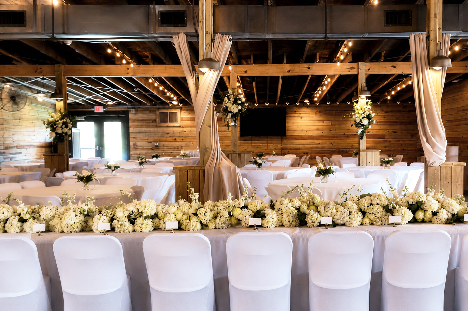 A decorated event space with round tables, white chairs, and floral centerpieces in front of a wooden wall and ceiling. String lights hang overhead, and beige curtains are tied back on wooden posts. There is a floral arrangement at the front on a lon