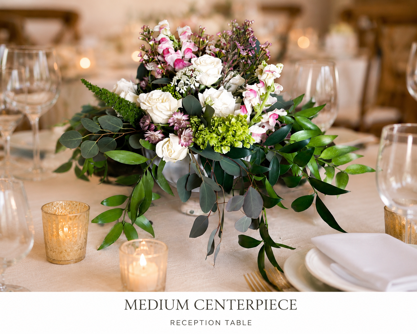 A floral centerpiece with white roses, pink and white orchids, and various greenery on a reception table, with candles and glassware around it.