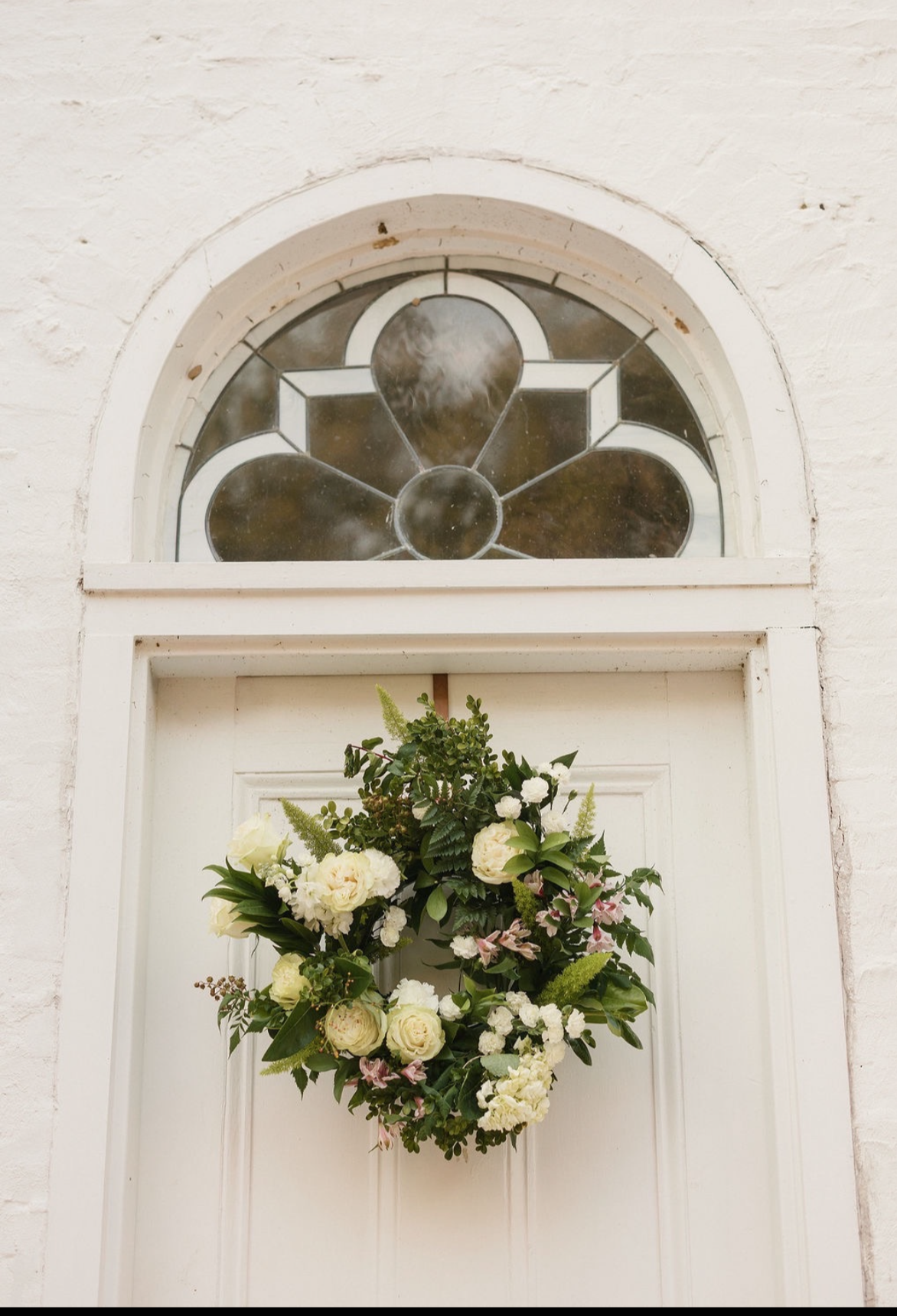 Wedding floral arrangement hanging on a white door with a stained glass window above it.