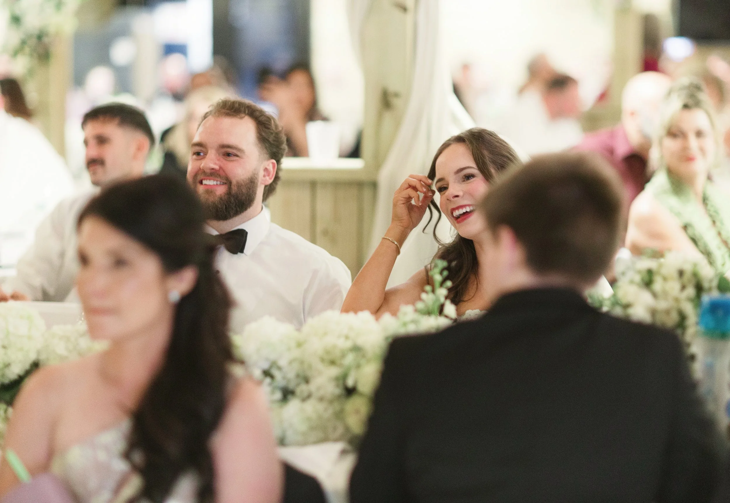 People sitting at tables with flower arrangements, smiling and enjoying an event in a decorated venue.